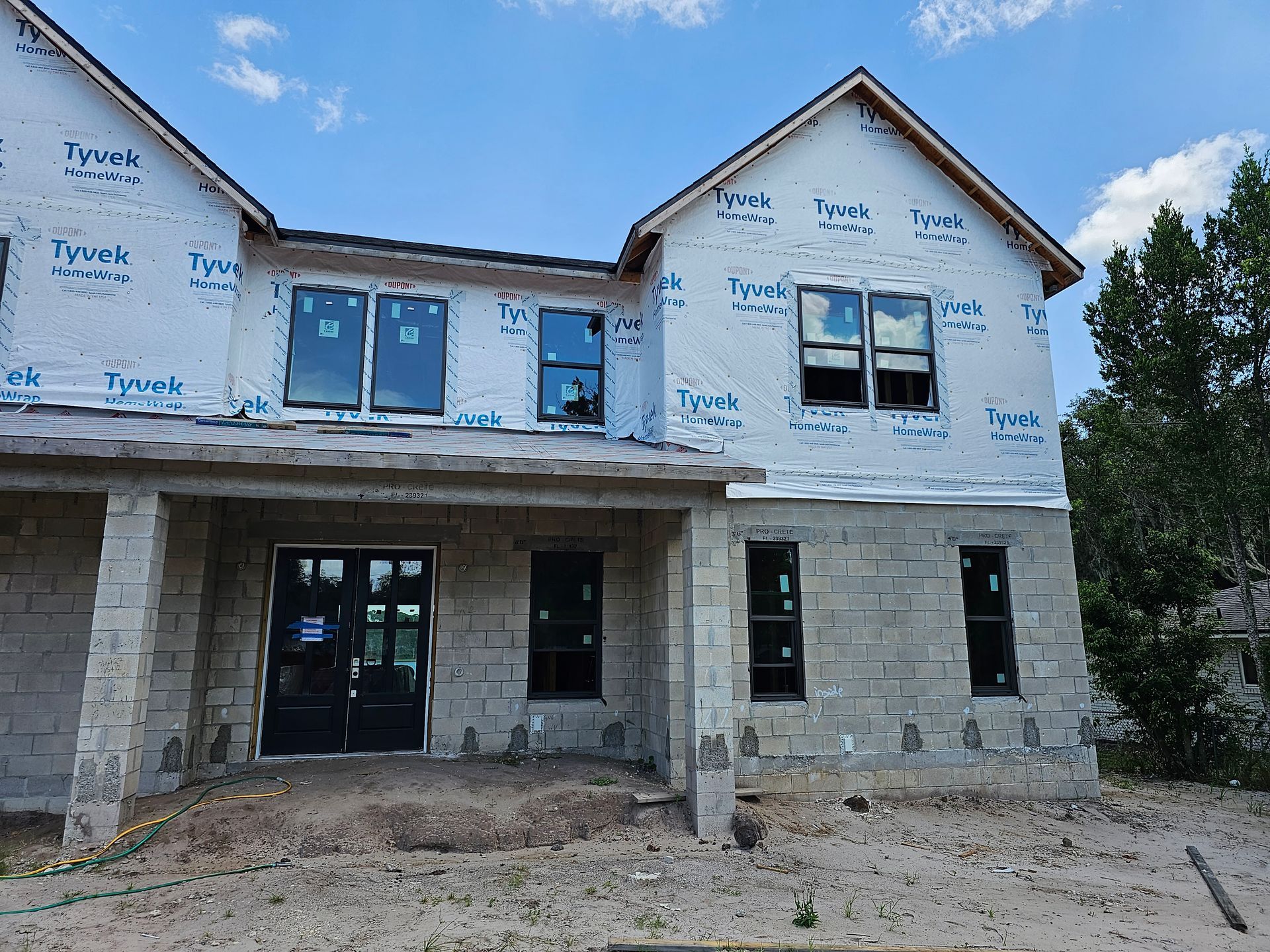 Two-story house under construction with white wrap, stone lower facade, and blue-trimmed windows and doors