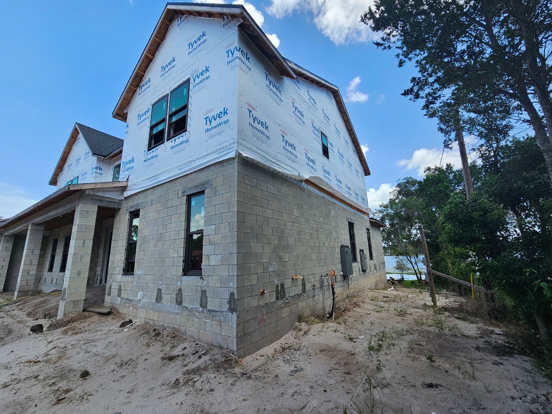 Partially built two-story house with exposed stone walls and white siding in a sandy wooded area