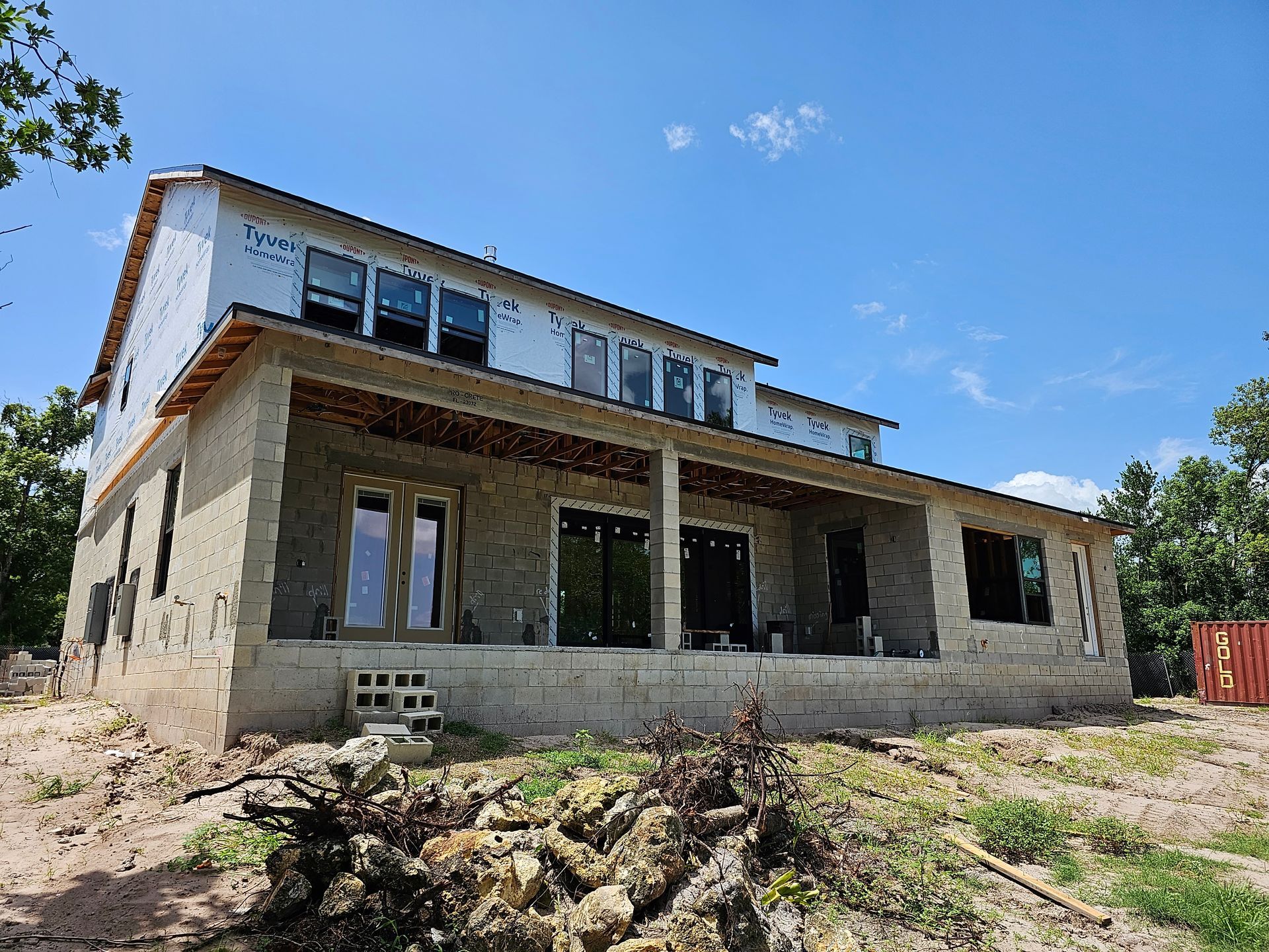 Partially built stone house with a covered porch on a sunny, grassy lot under a blue sky