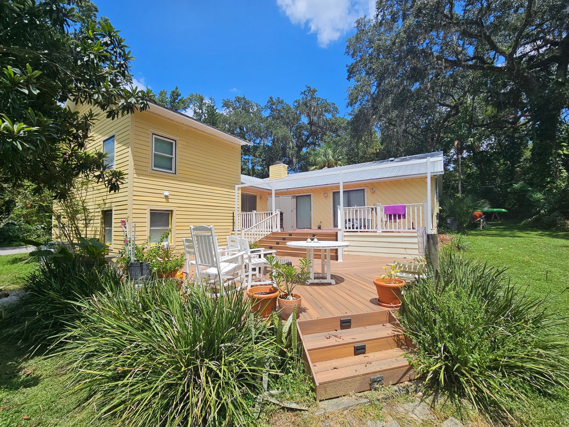 Yellow house with white porch, wooden steps, and patio furniture surrounded by lush greenery