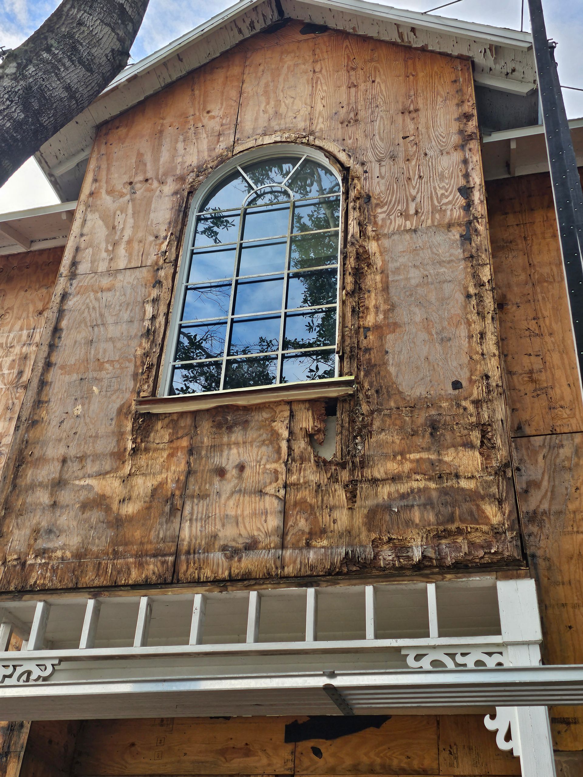 Weathered wooden building facade with a tall arched window and peeling brown exterior under a roof overhang