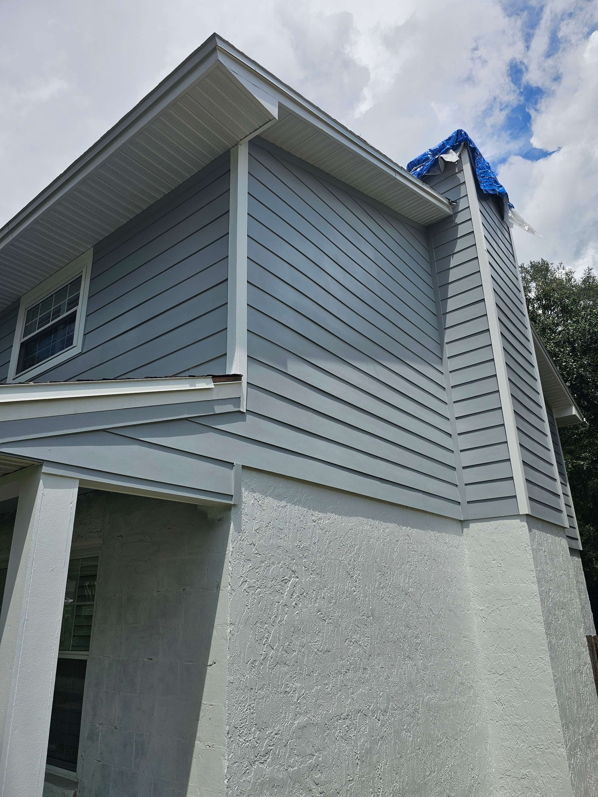 Exterior corner of a gray house with siding, a textured lower wall, and a blue tarp on the roofline