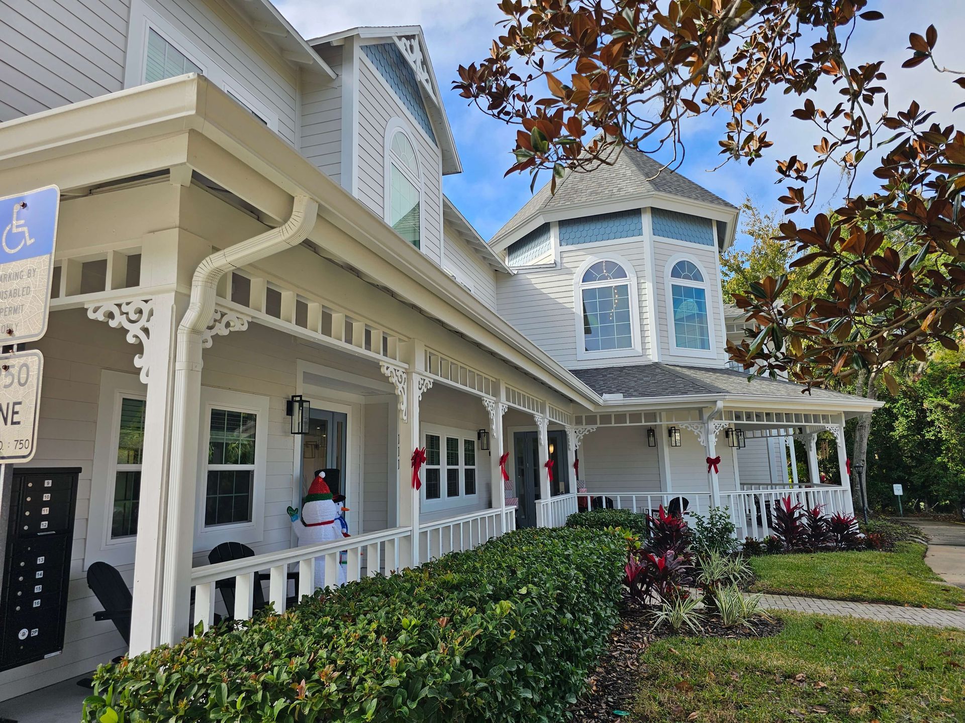 Victorian-style white house with a porch, blue shutters, and shrubs under a partly cloudy sky