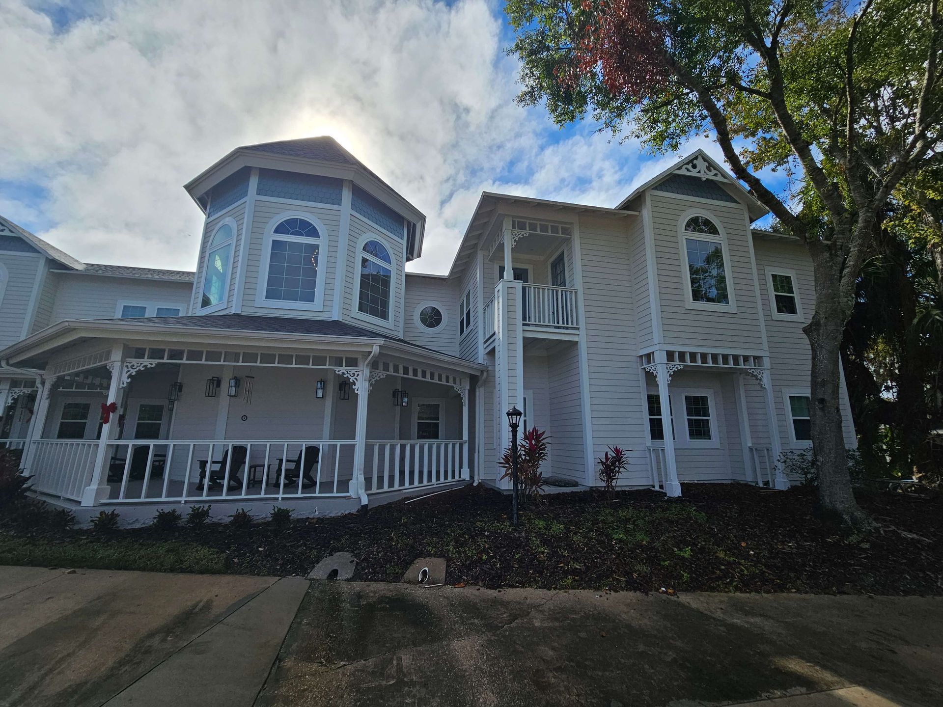 White Victorian-style house with wraparound porch, bay windows, and dark roof under a partly cloudy sky