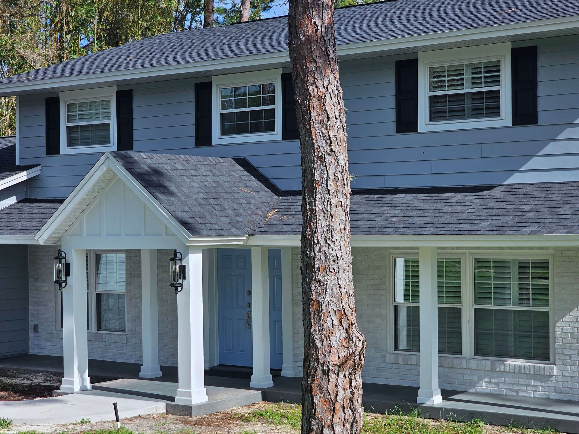 Gray two-story house with white trim, black shutters, and a small front porch under tall trees