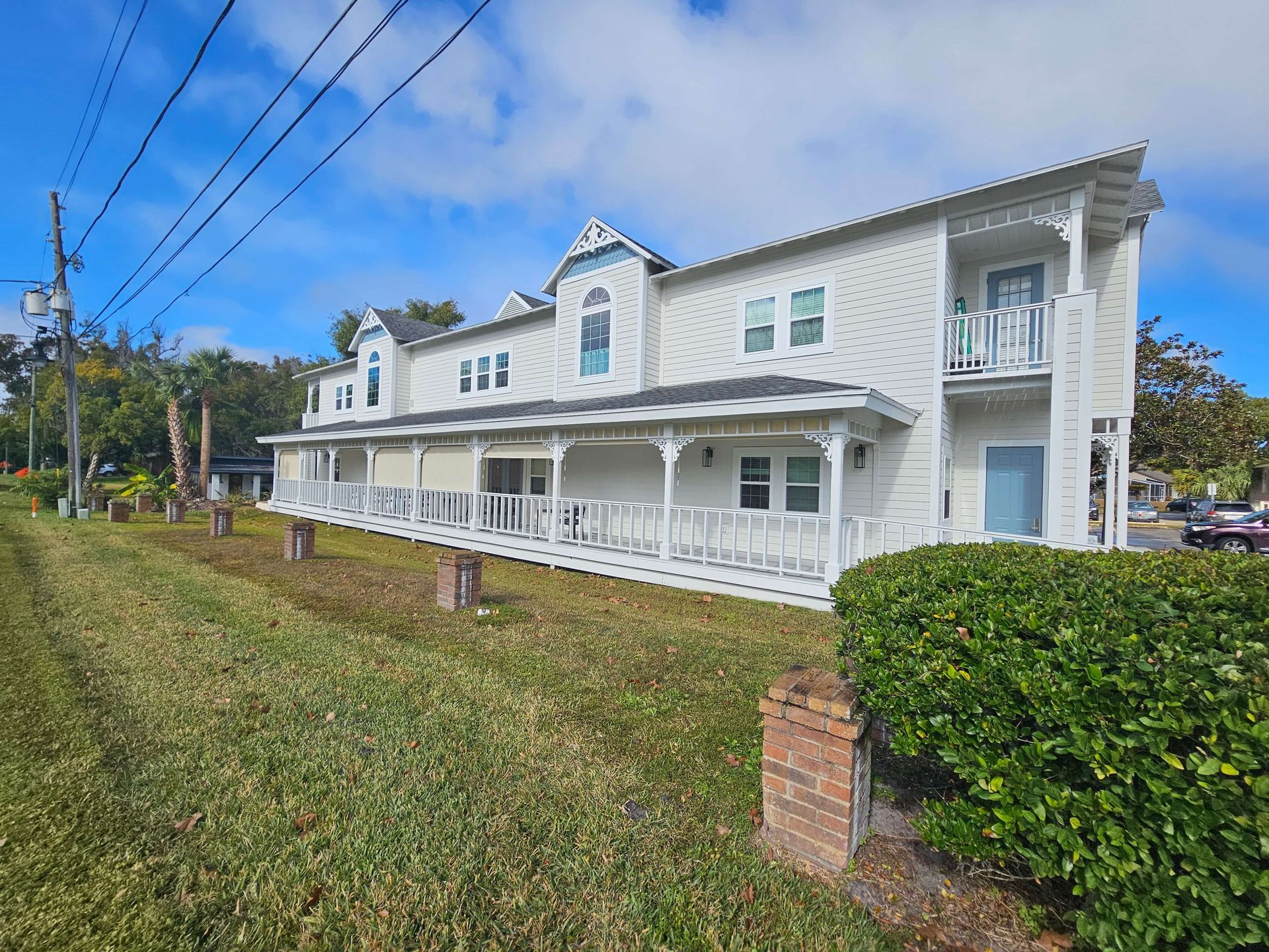 White two-story house with wraparound porch beside a grassy roadside under a blue sky