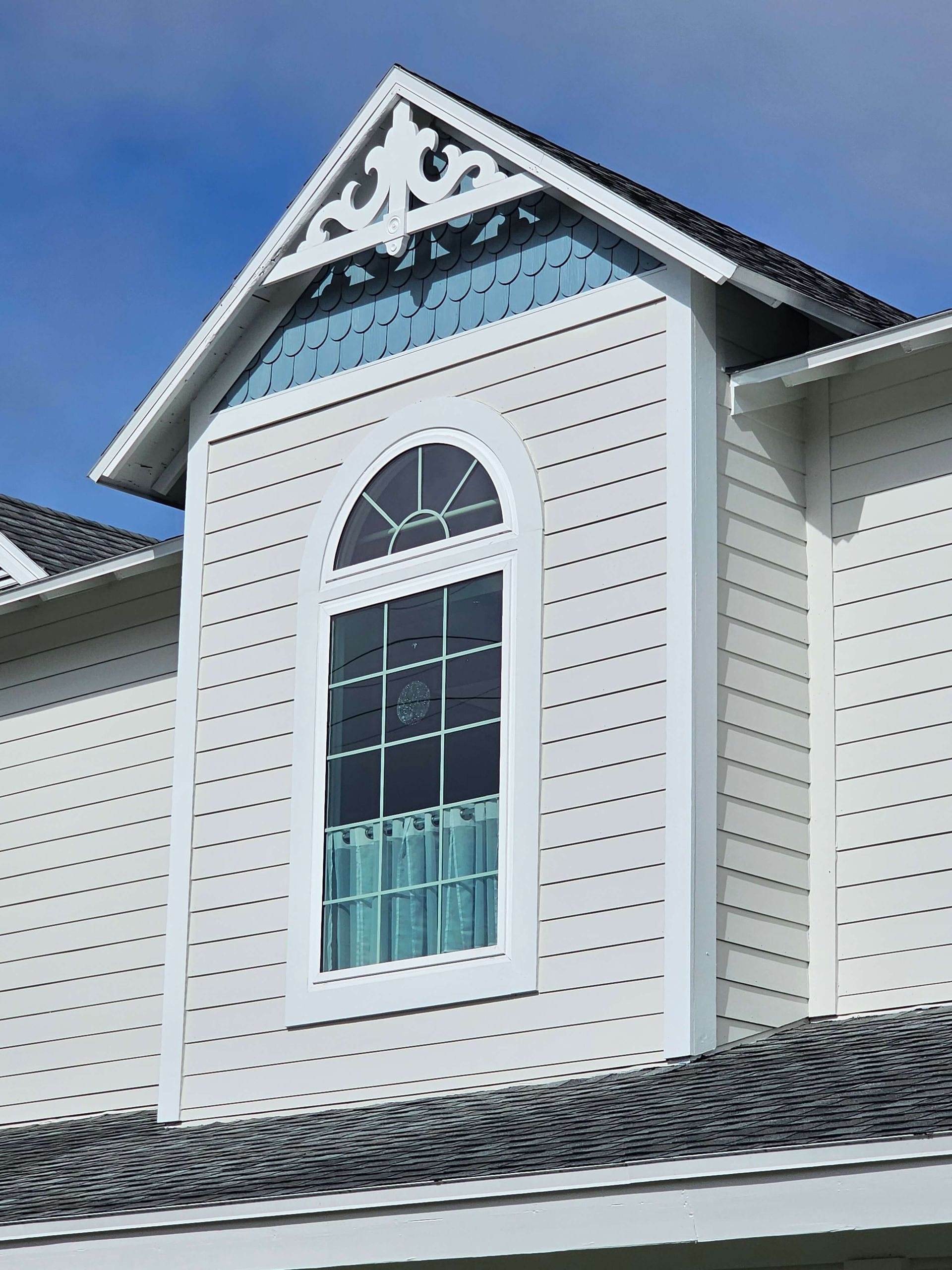 White house dormer with arched window, blue curtains, and decorative trim under a blue sky