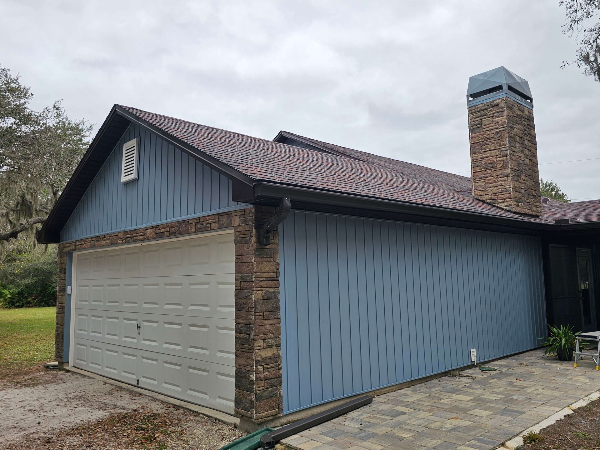 Blue-sided house with a garage, brick chimney, and driveway under an overcast sky