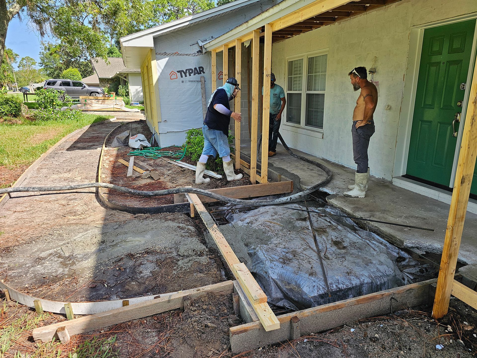 Workers building a porch frame beside a white house with yellow posts and green doors