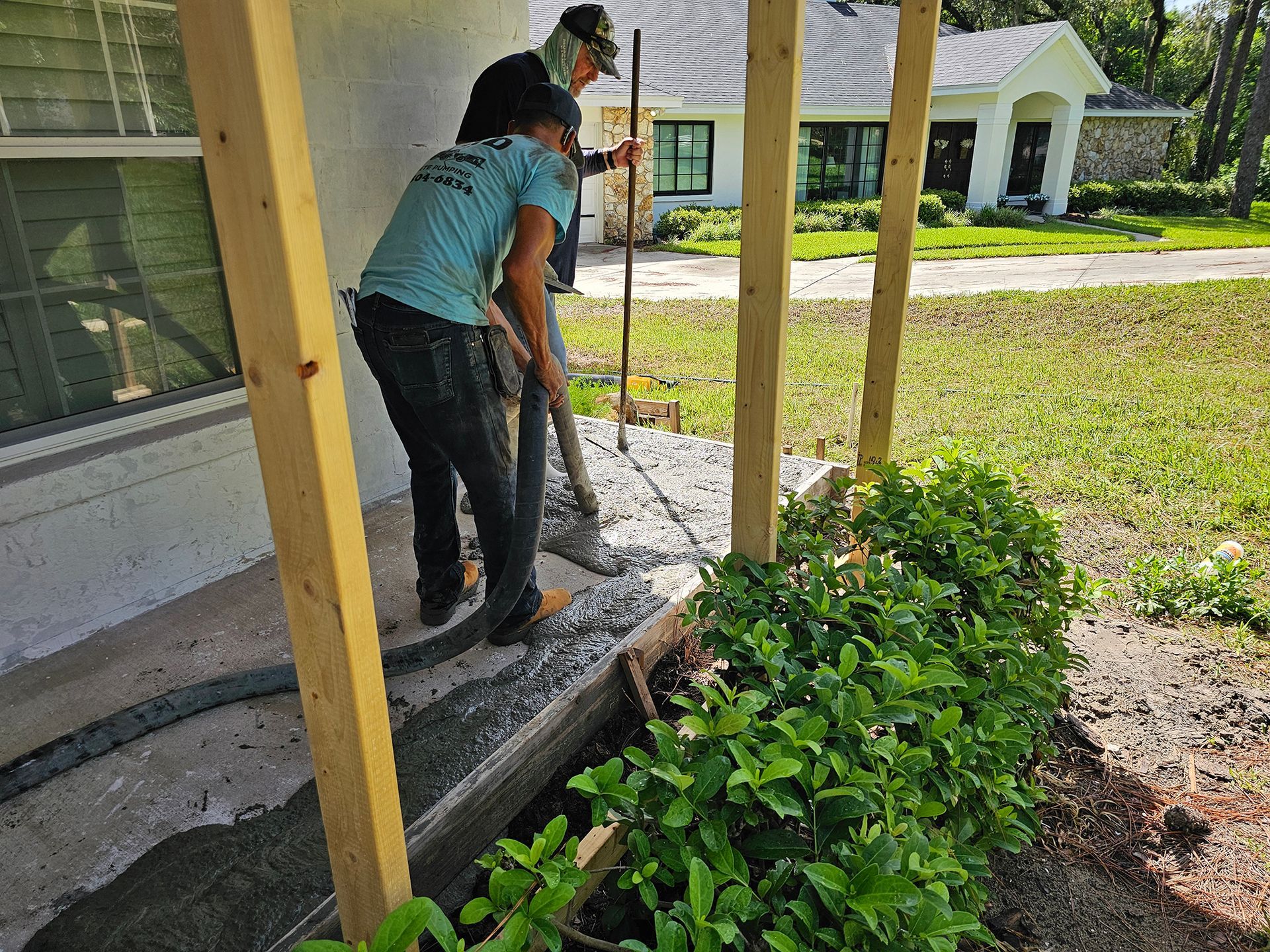 Person painting a porch post beside a house, with green shrubs and a sunny yard.