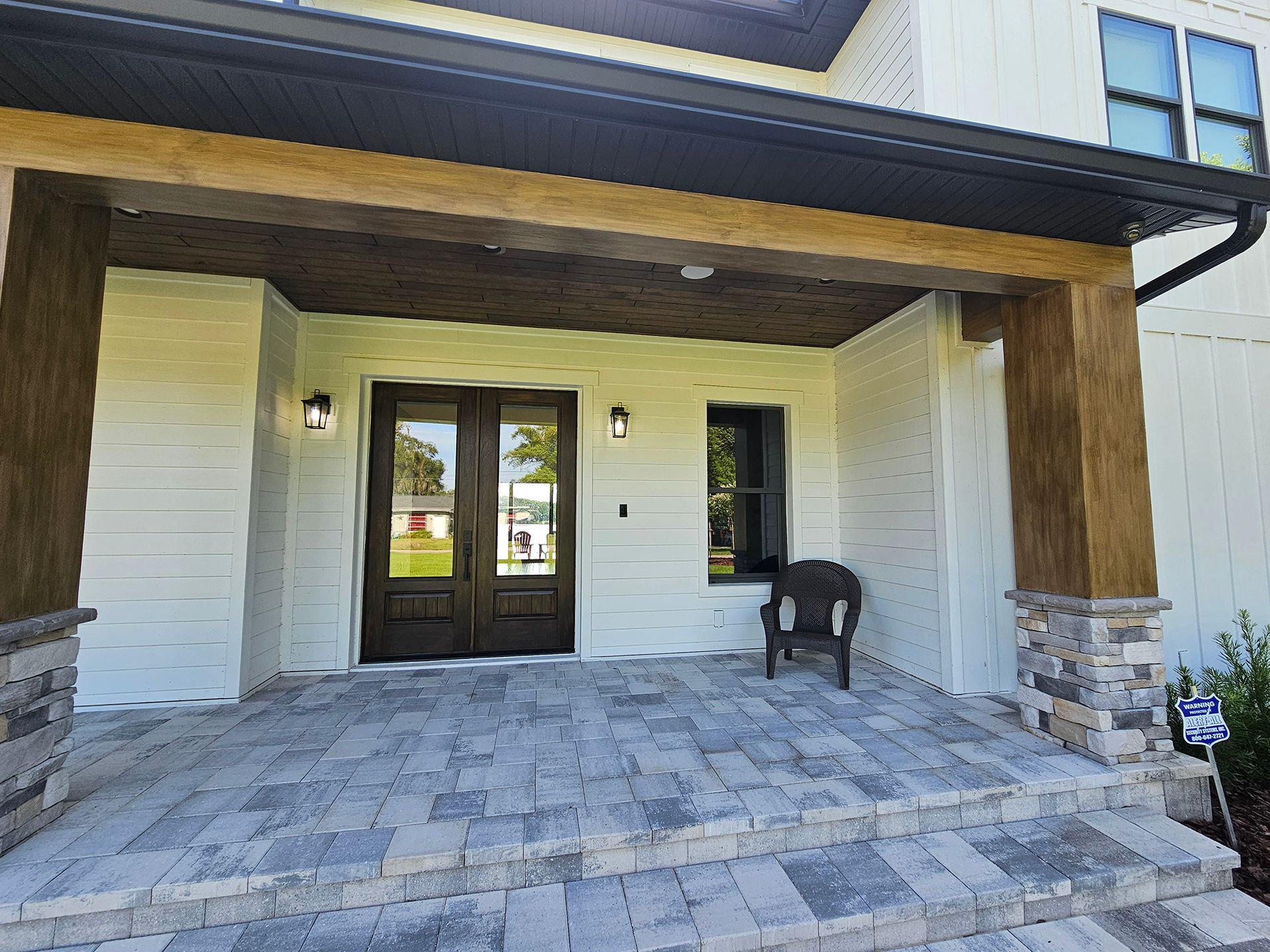 Covered front porch with stone pavers, white siding, dark trim, and a chair by the front door.