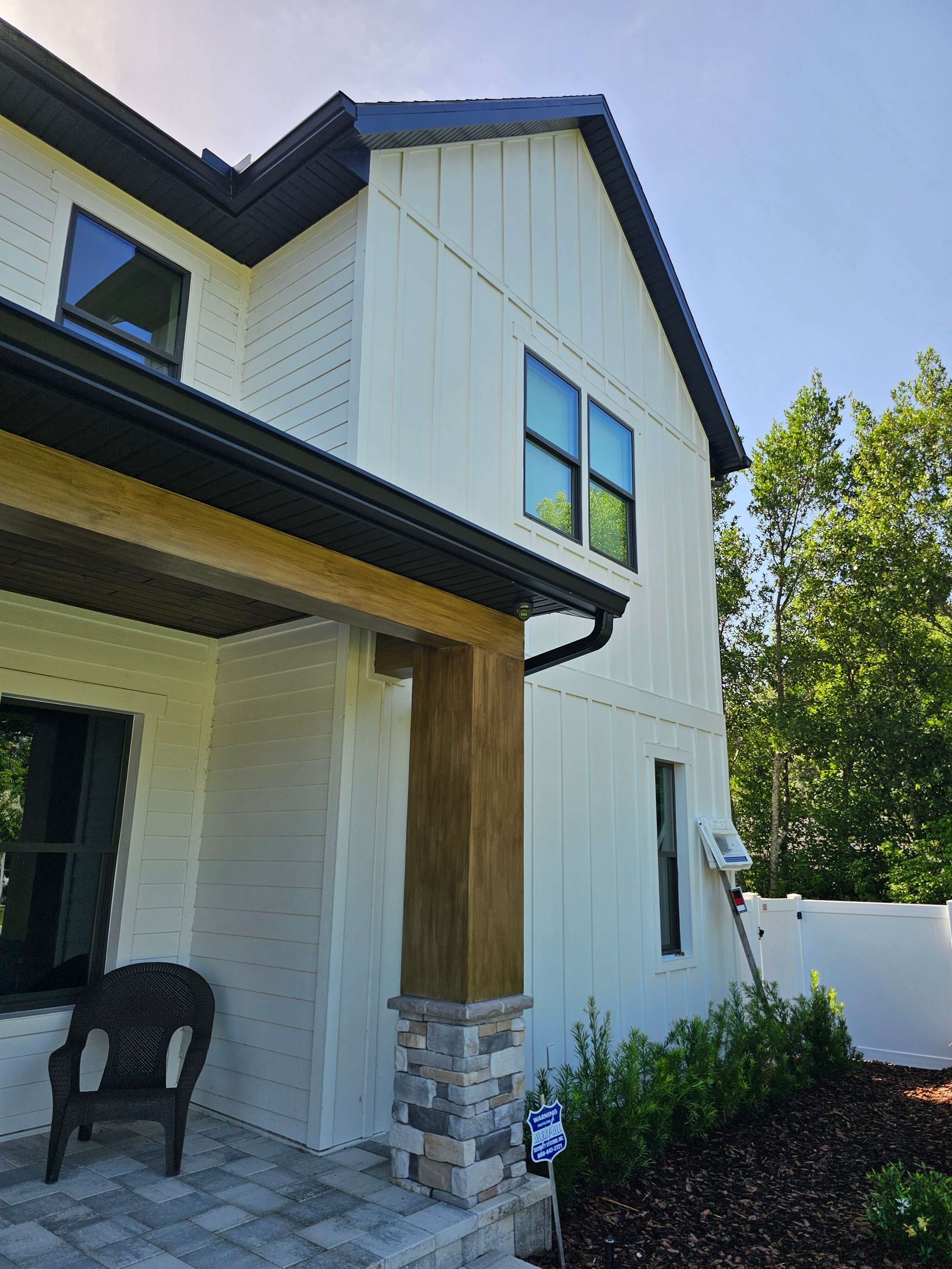 White two-story house exterior with black trim, stone column, and front porch chair under bright sunlight
