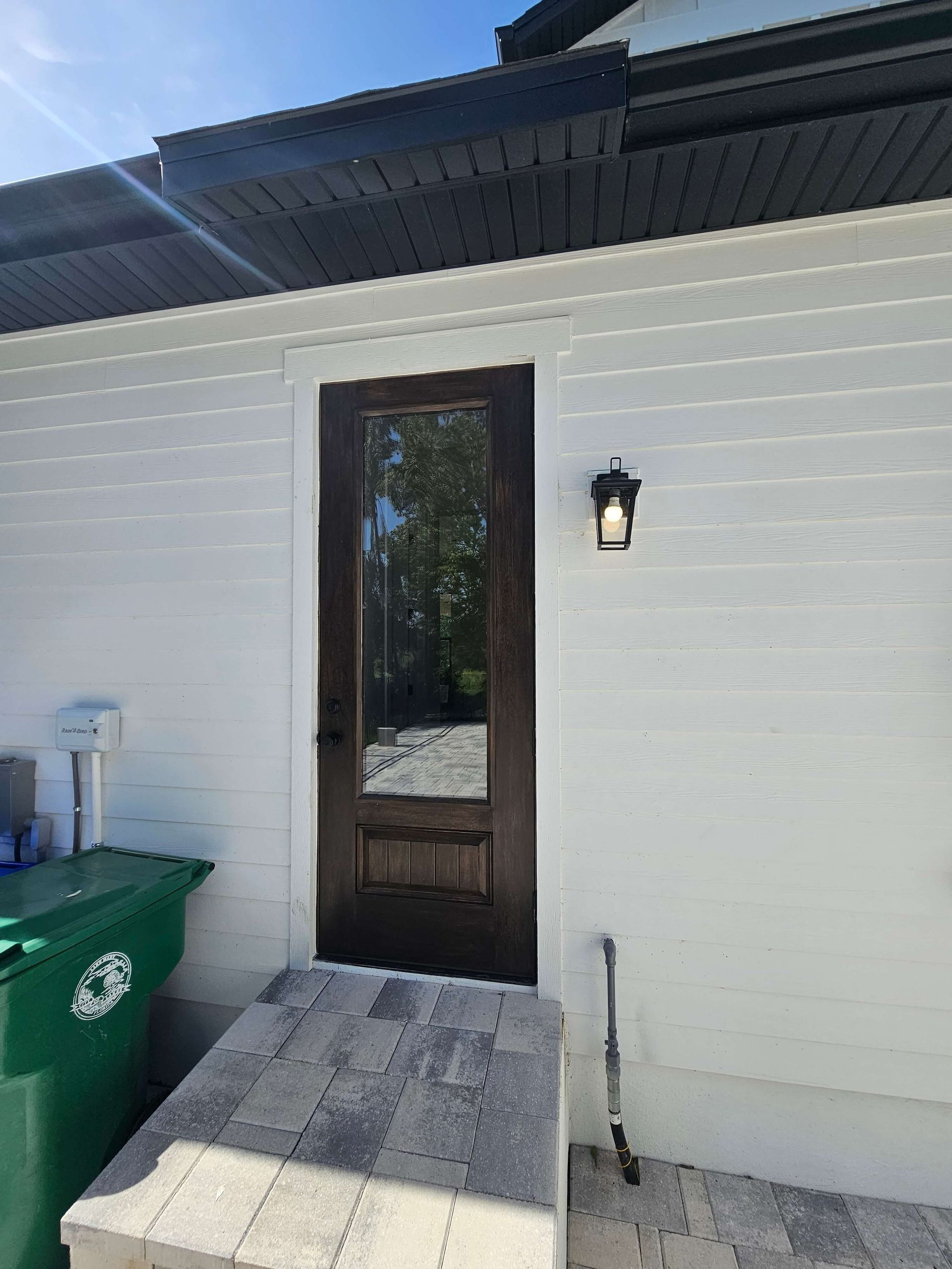 White house exterior with a dark wooden door, wall light, and green trash bin beside a concrete step.