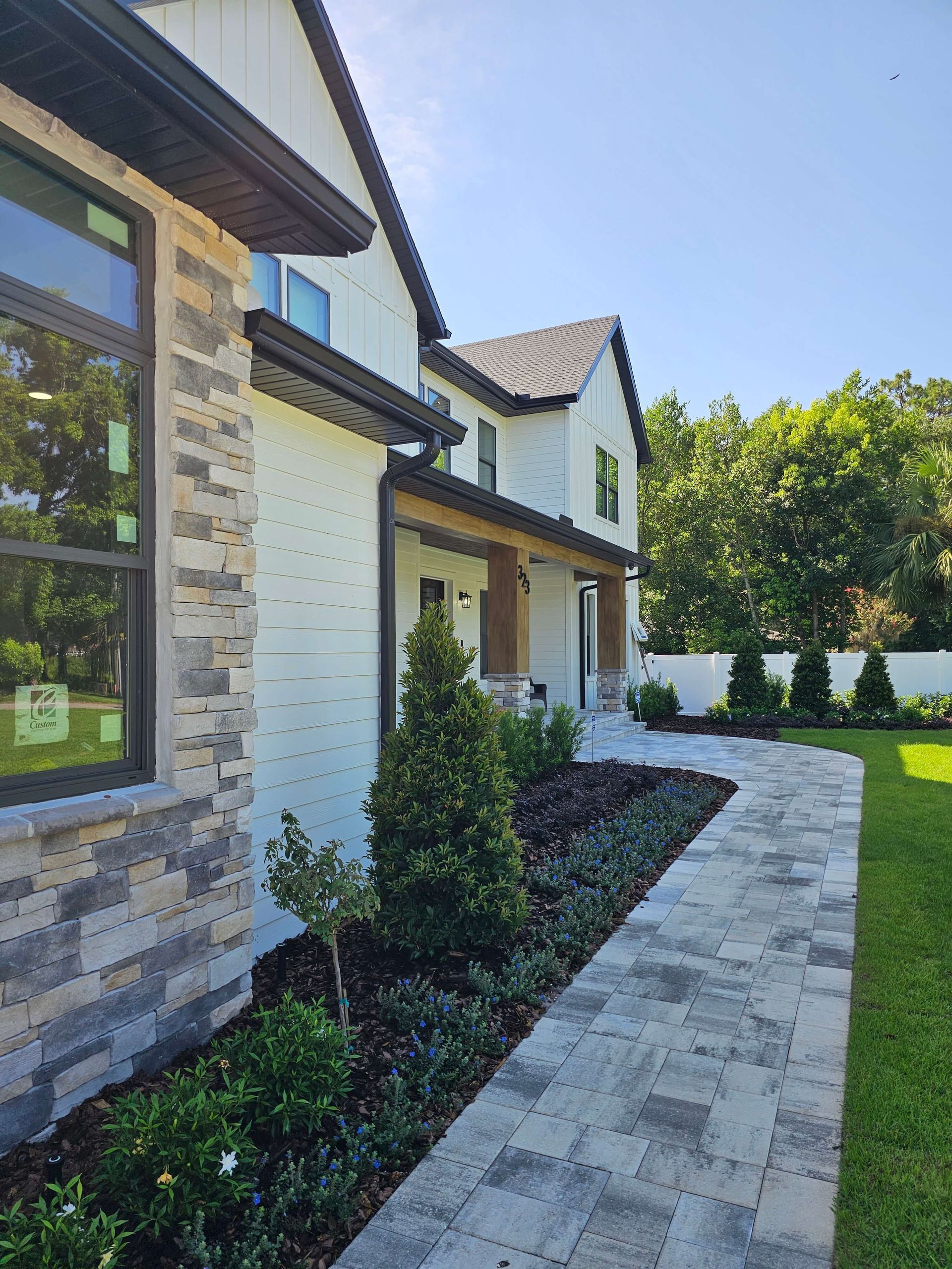 Modern house exterior with white brick, stone accents, and a curved walkway beside landscaped shrubs.