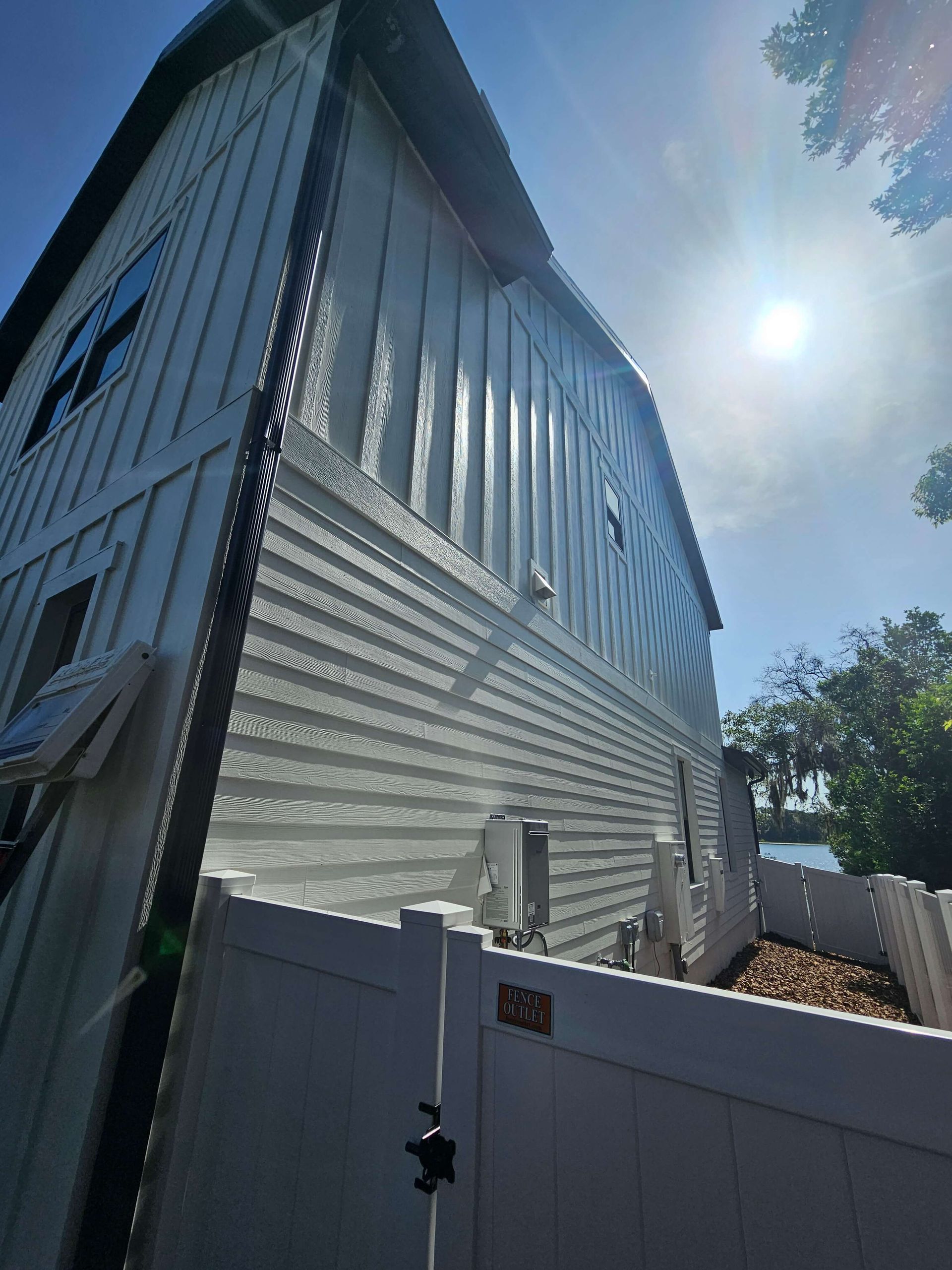 Tall silver-sided house seen from below behind a white fence under a bright sun and blue sky