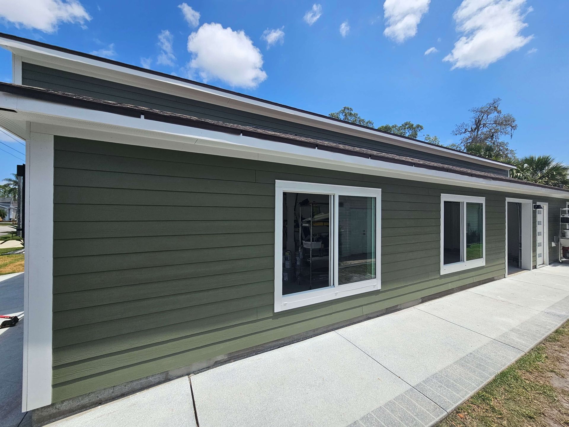 Modern green building exterior with white trim, large windows, and a concrete walkway under a blue sky