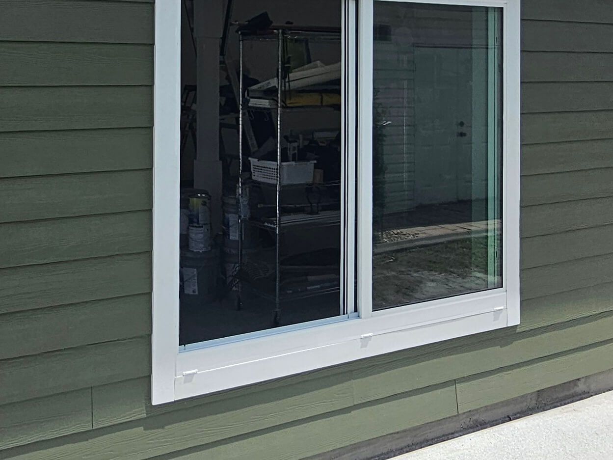 Sliding window with white trim on green house siding, showing interior shelves through the glass.