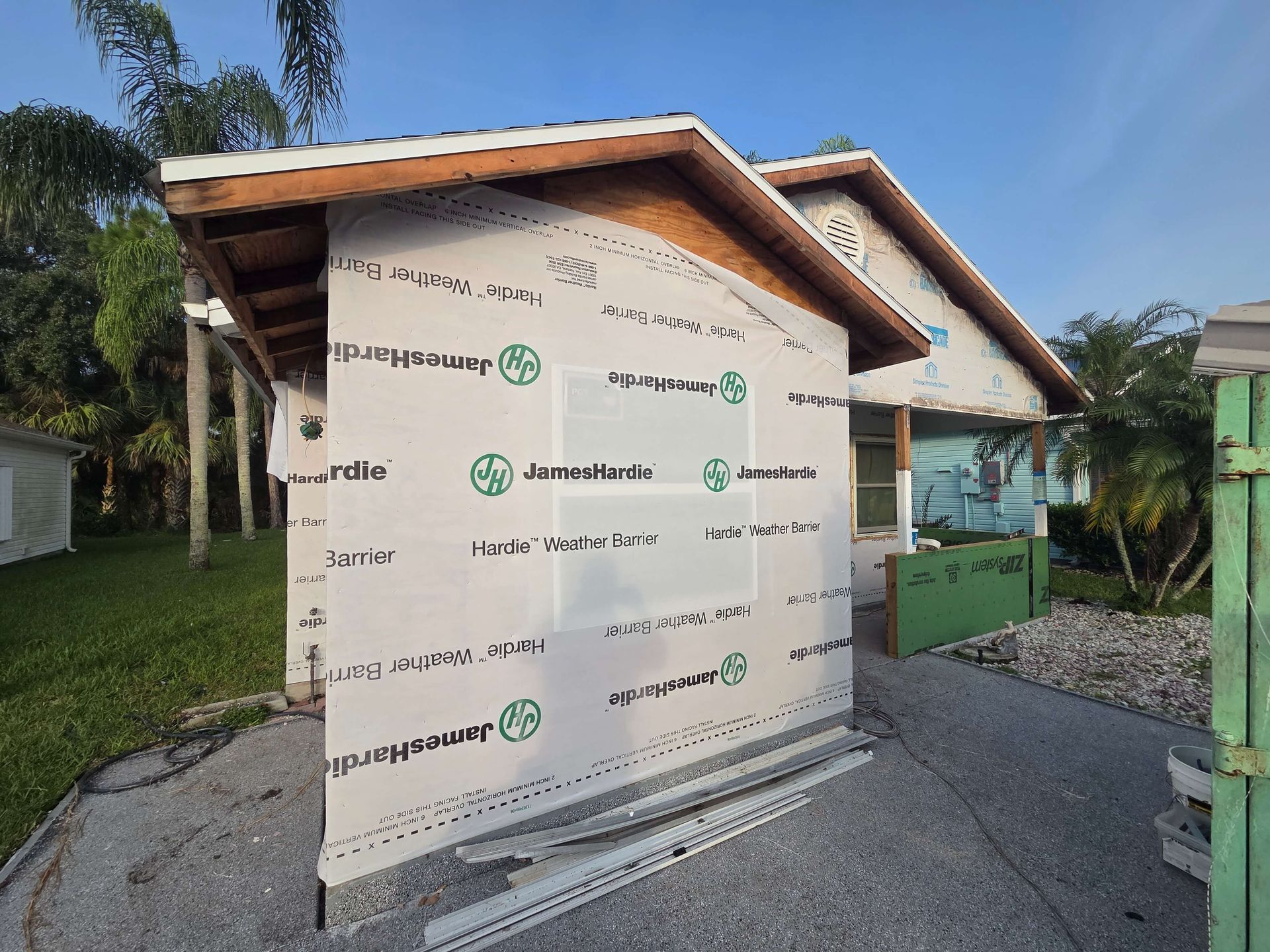 Small unfinished house exterior with wrapped siding, gravel driveway, and trees beside it