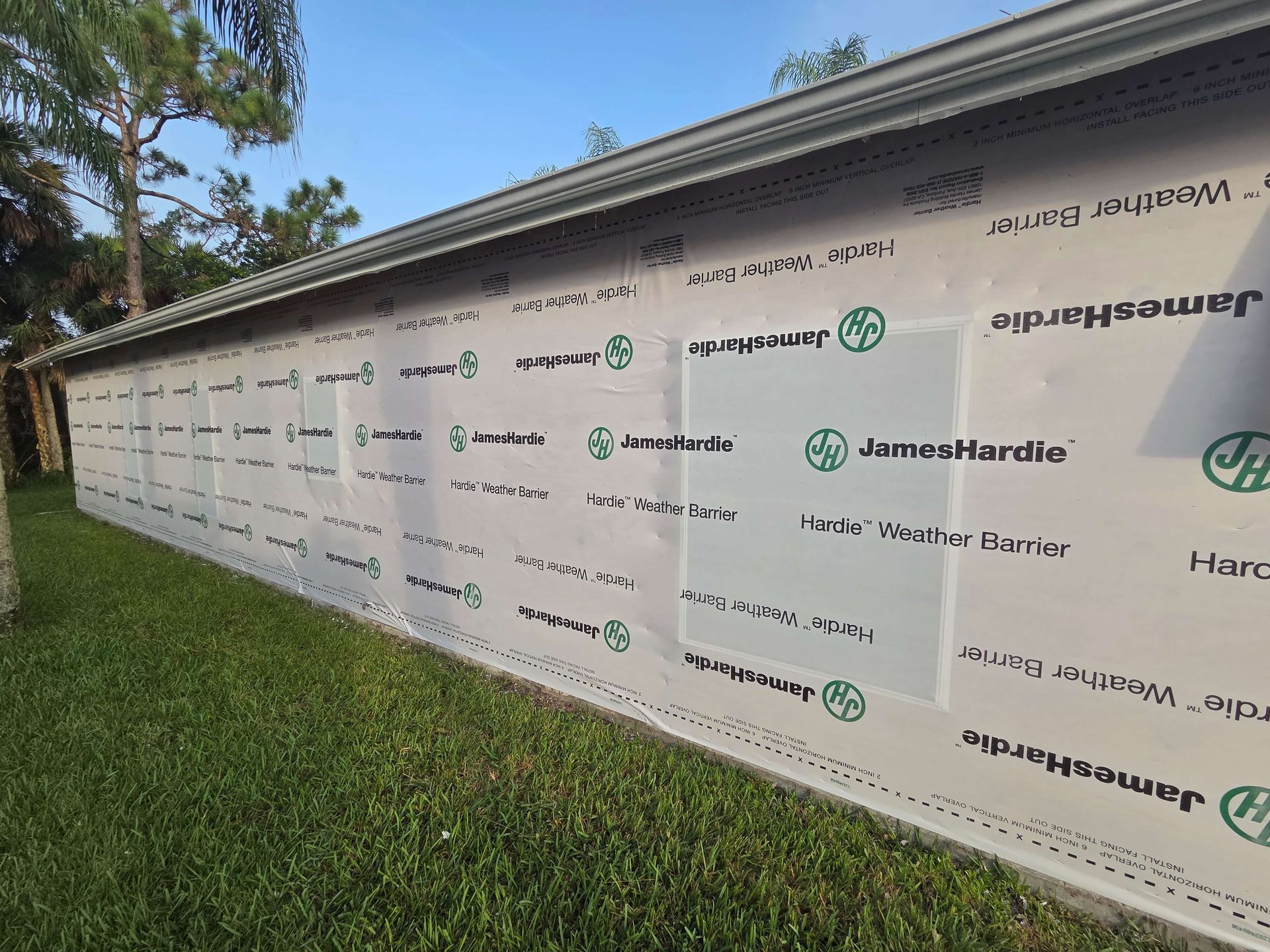 Construction wrap with repeated logos on a long building wall beside green grass and trees