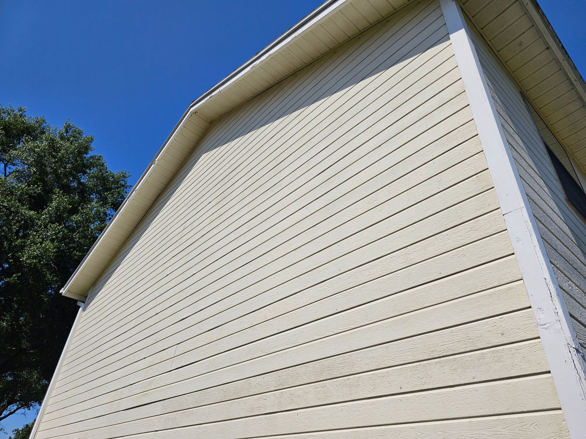 Close-up of beige house siding and white trim under a roofline against a blue sky.