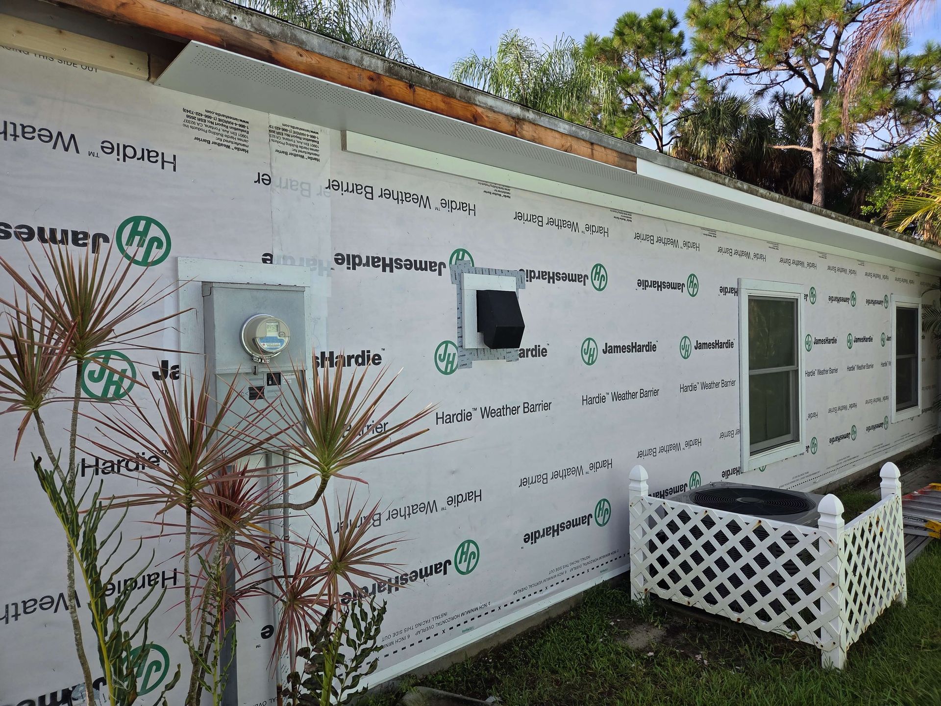 House exterior wrapped in white Tyvek, with windows, utility meter, and a small lattice planter on the right.