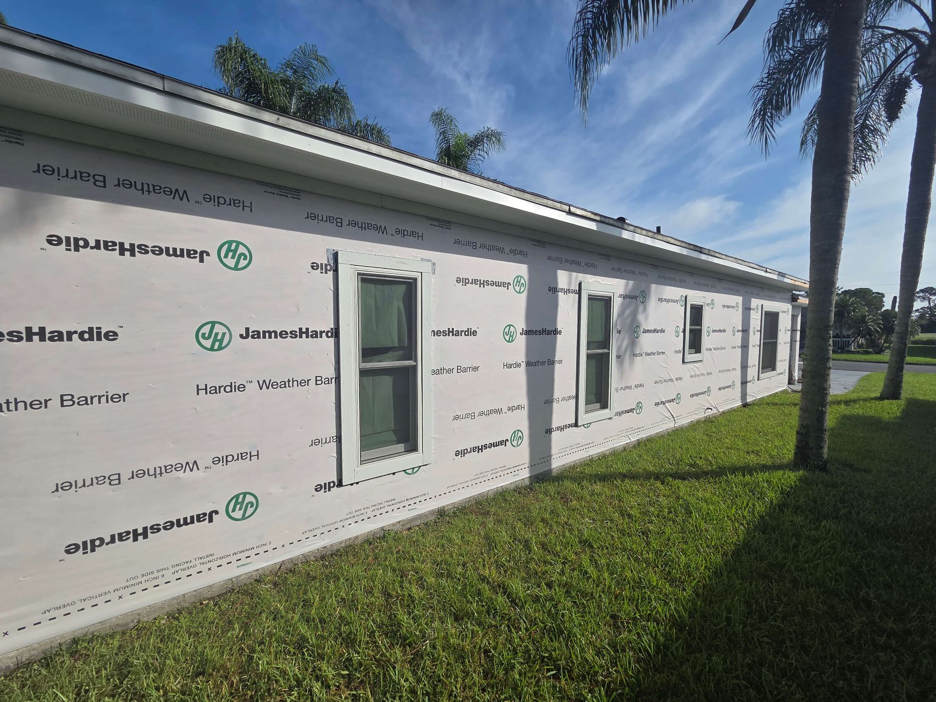 White modular building with windows, wrapped in house wrap, beside a grassy lawn and palm trees.