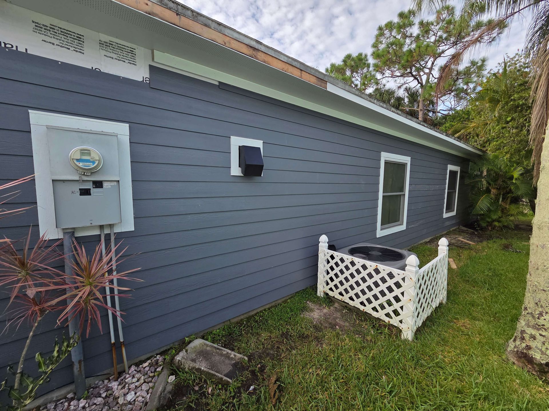 Blue-gray house side with utility meter, window, and small white fenced garden bed by a tree
