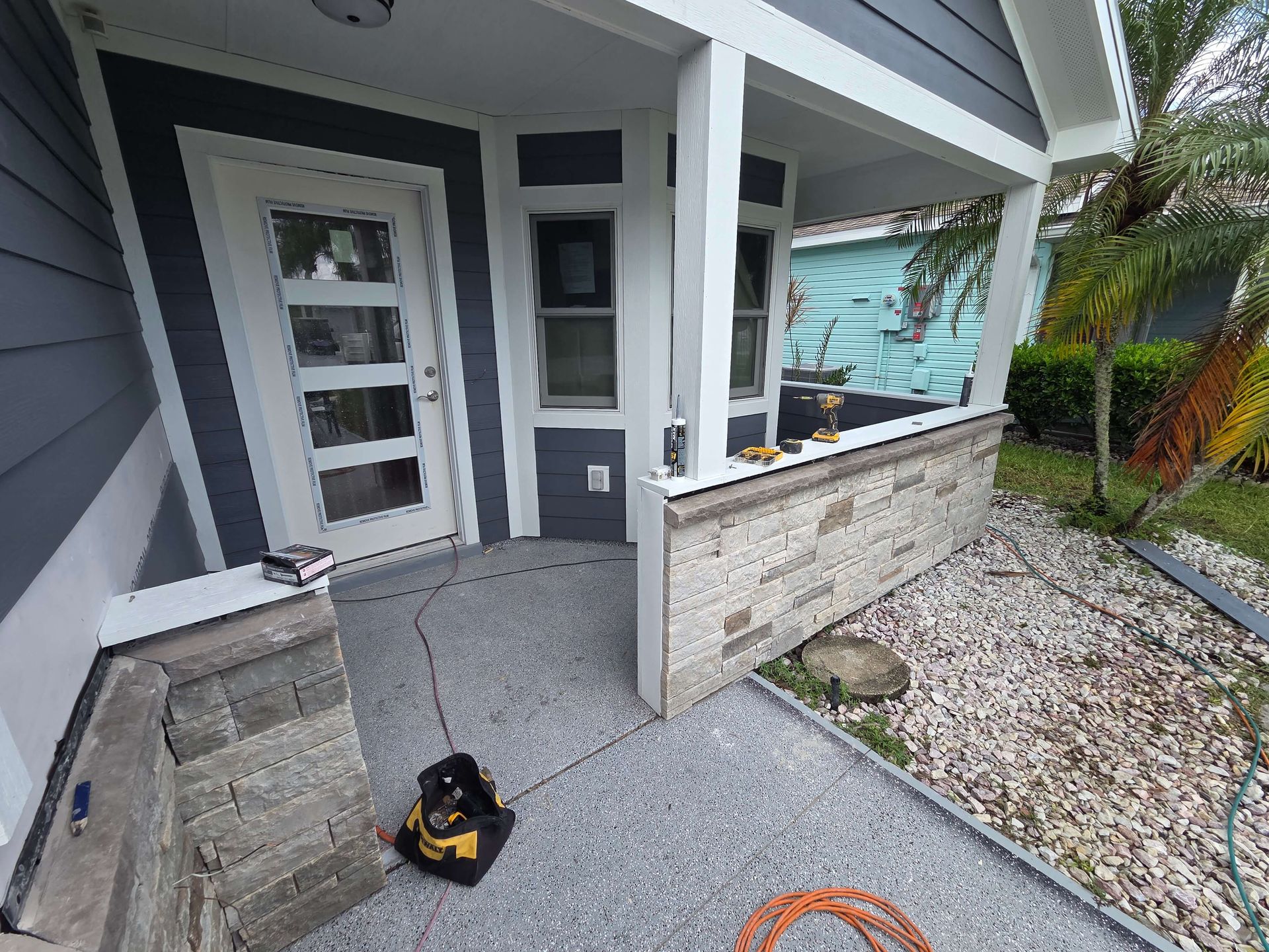 Front porch with gray siding, white door, stone half-wall, and a black-and-yellow vacuum on the concrete floor.