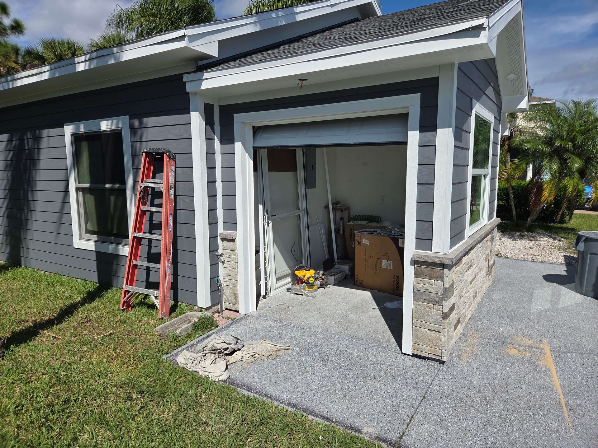 Grey house exterior with open garage, red ladder, and stone trim beside a gravel driveway