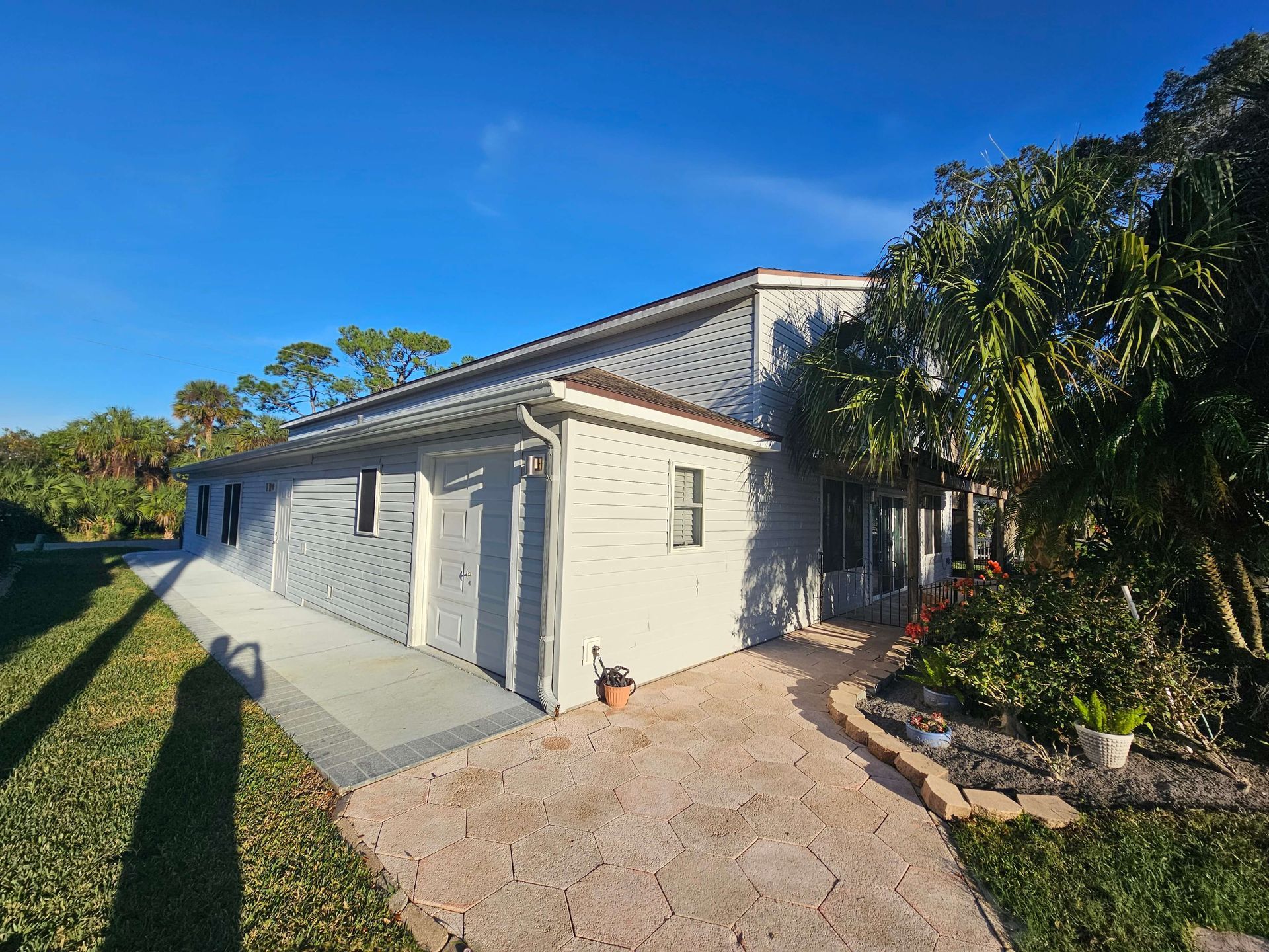 Single-story light gray house with a tiled roof, front walkway, and palm trees in bright sunlight
