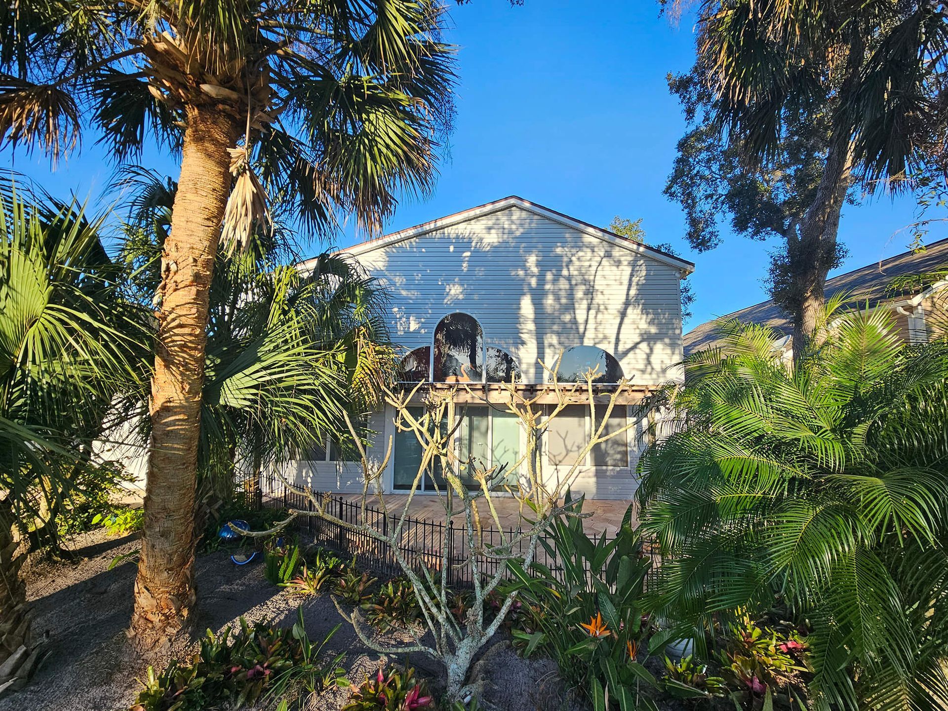 White house with a peaked roof behind palm trees and lush tropical plants under a blue sky
