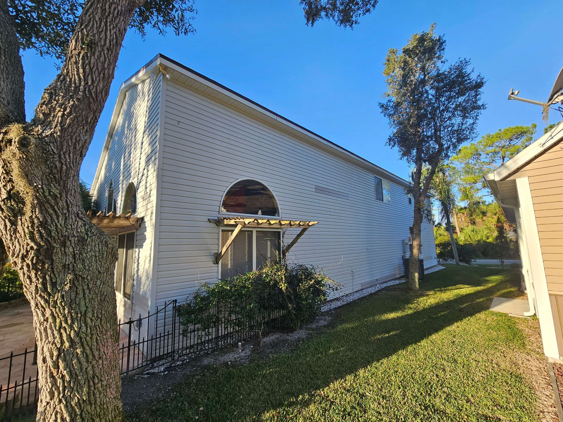 White house side wall with arched window, small awning, and trees in a sunny yard