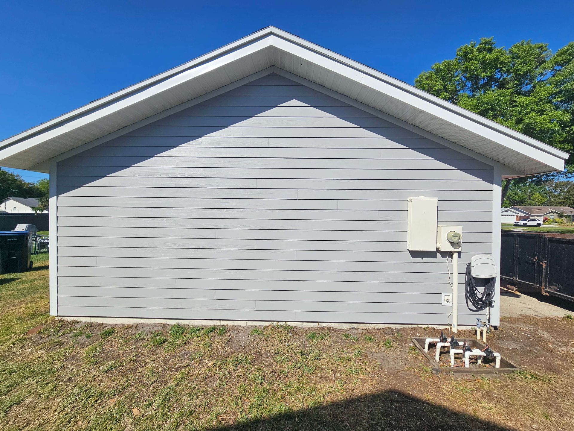 Gray house side wall with white trim, utility meter, and grassy yard under a blue sky