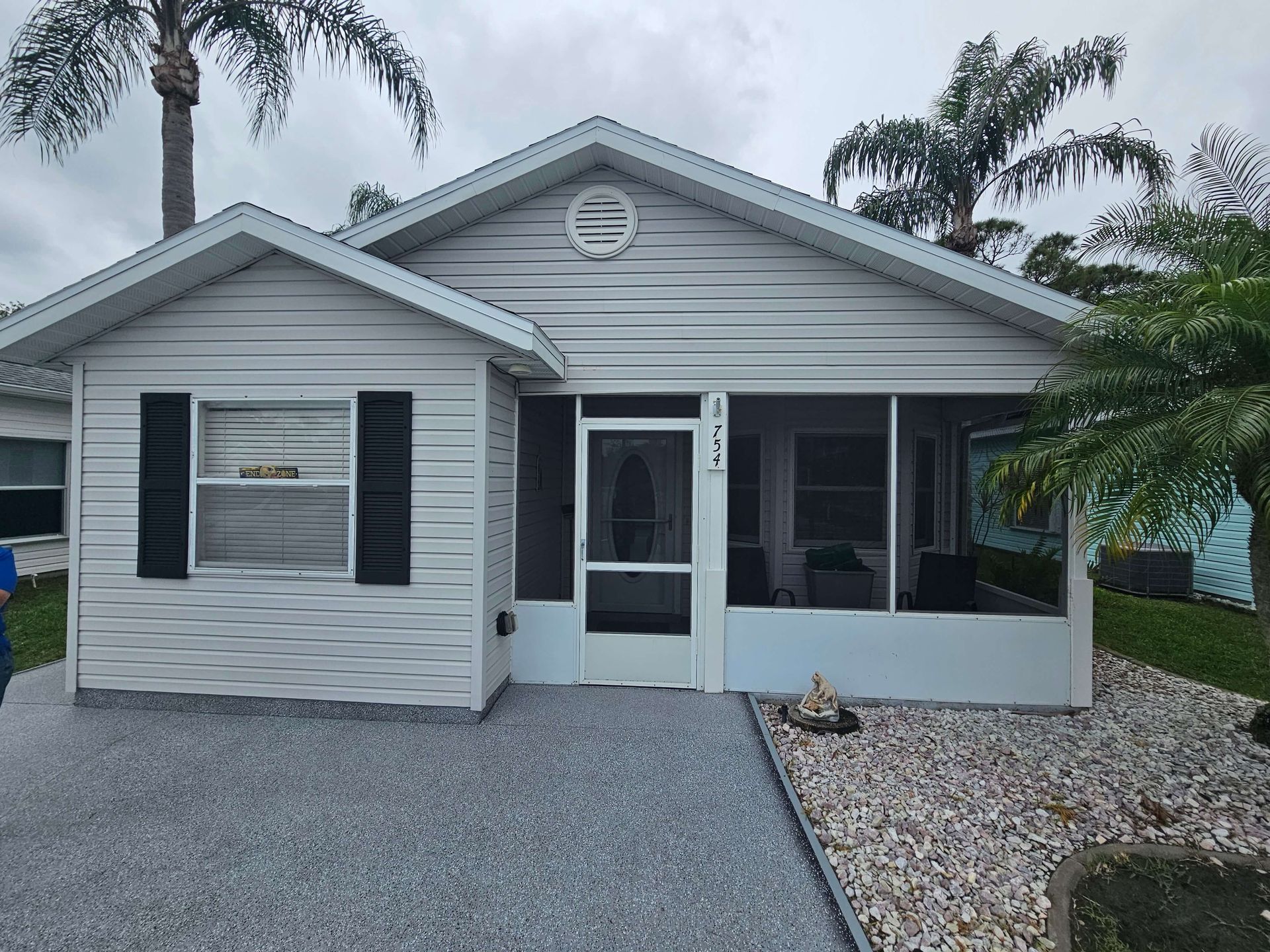 Small gray single-story house with black shutters, screened porch, and gravel yard under cloudy sky