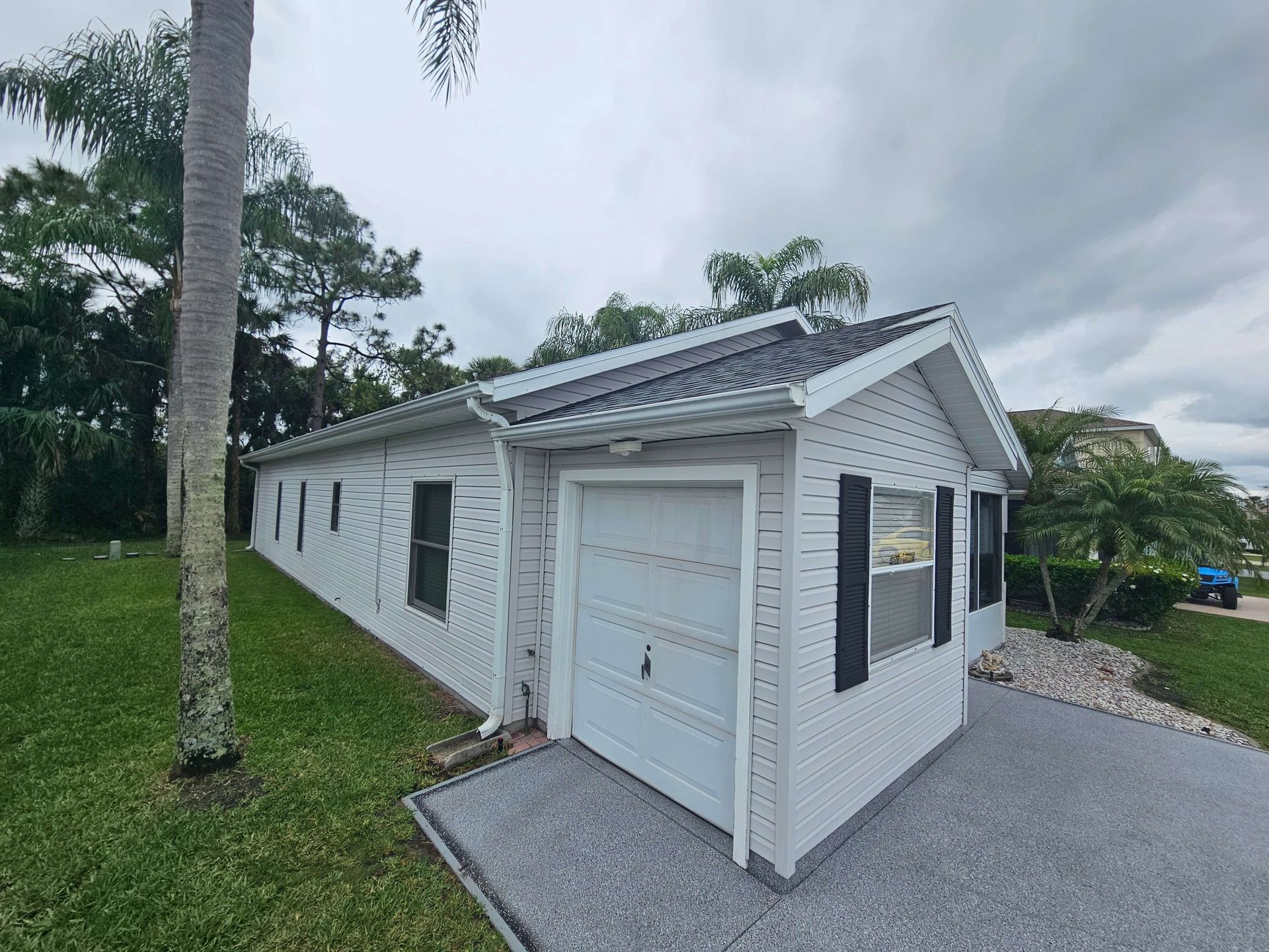 White garage with dark trim beside a gravel driveway and palm tree on a grassy hillside under cloudy sky