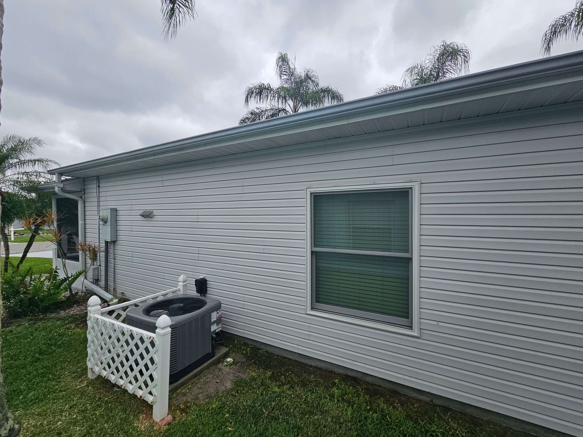 Side of a gray house with a window, small fenced AC unit, and cloudy sky overhead