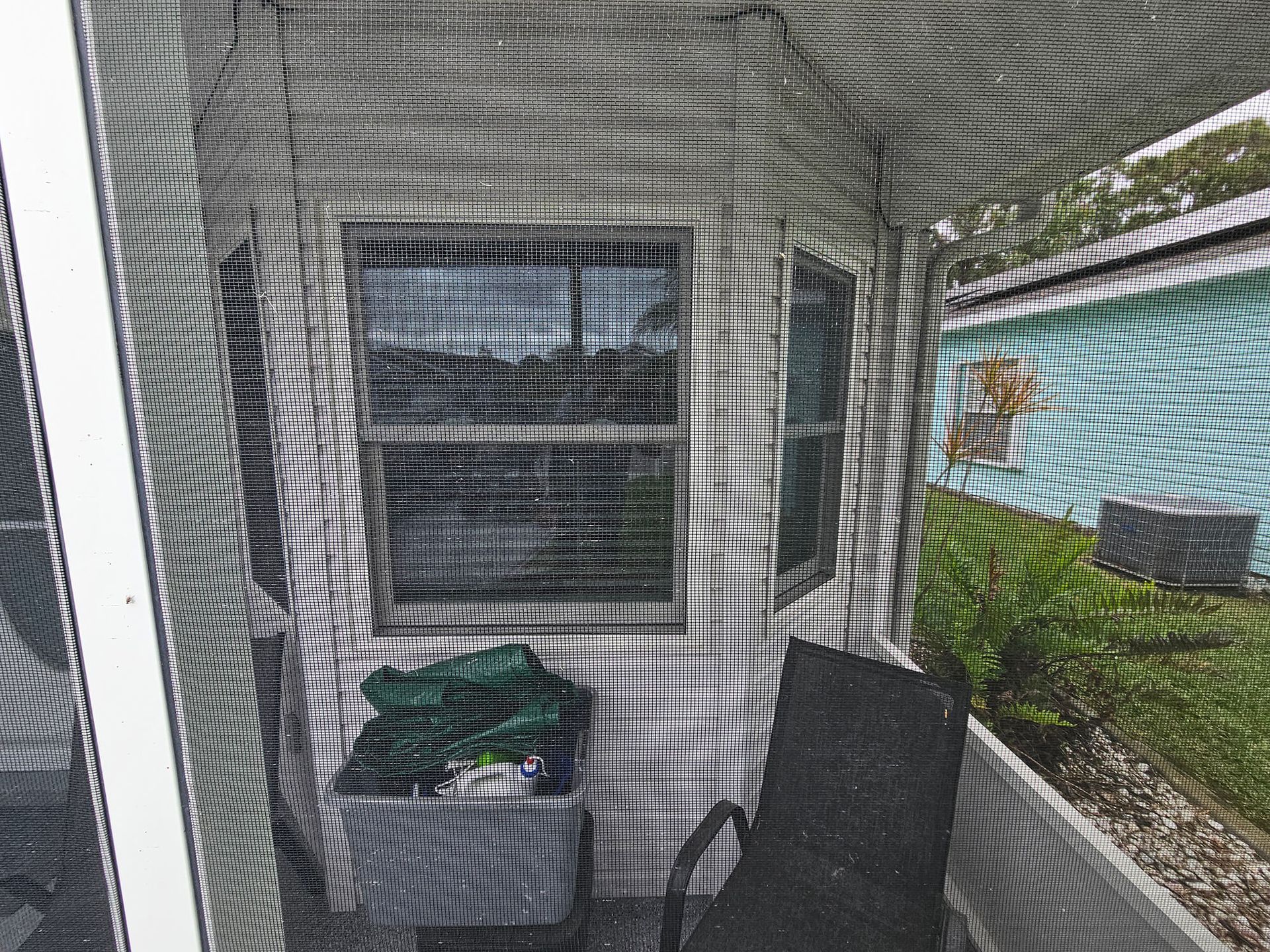 Screened porch with wicker chair, storage bin, and window air conditioner next to a grassy yard.