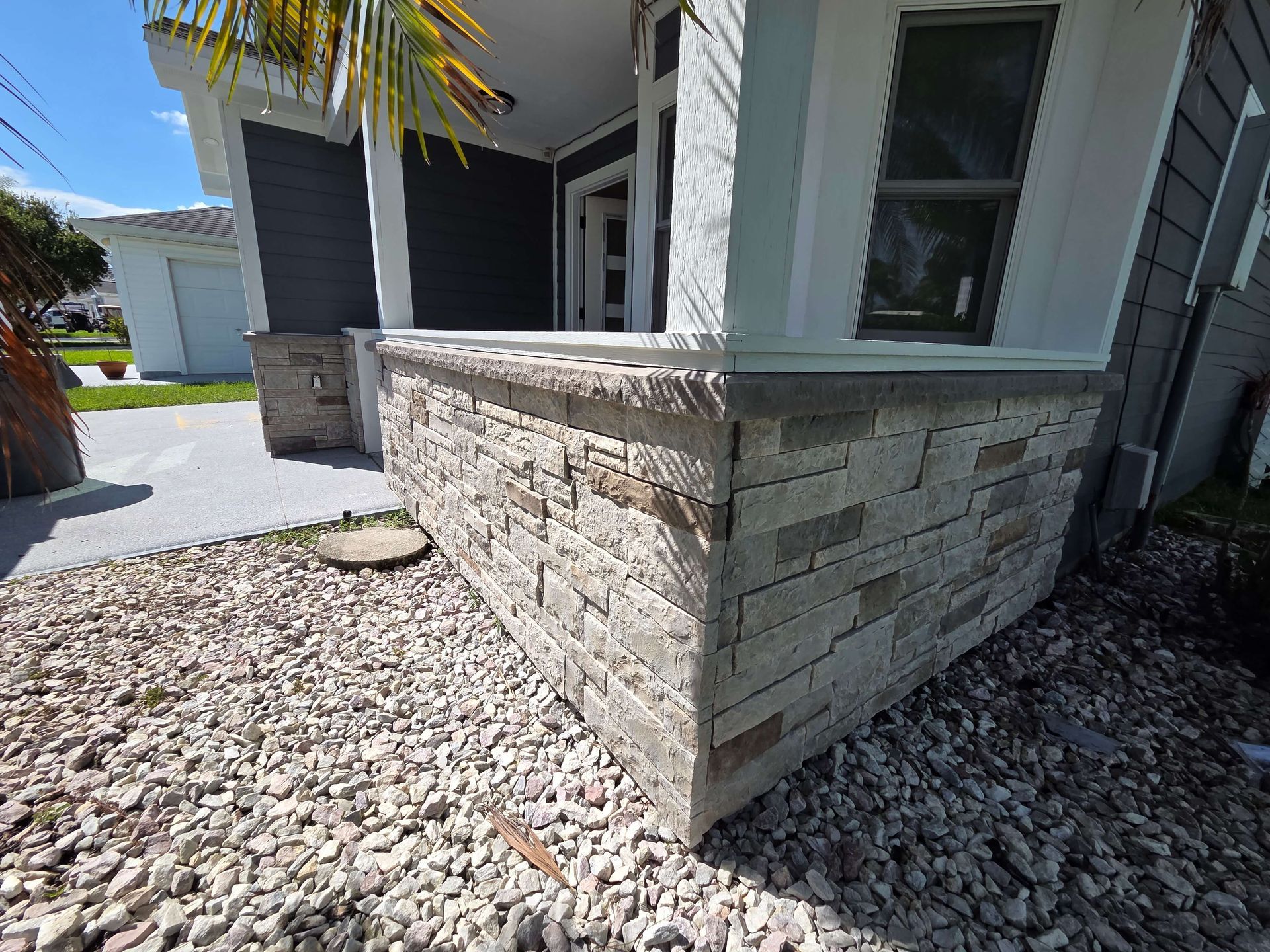 Stone planter wall beside a house with gravel landscaping and a walkway in bright sunlight