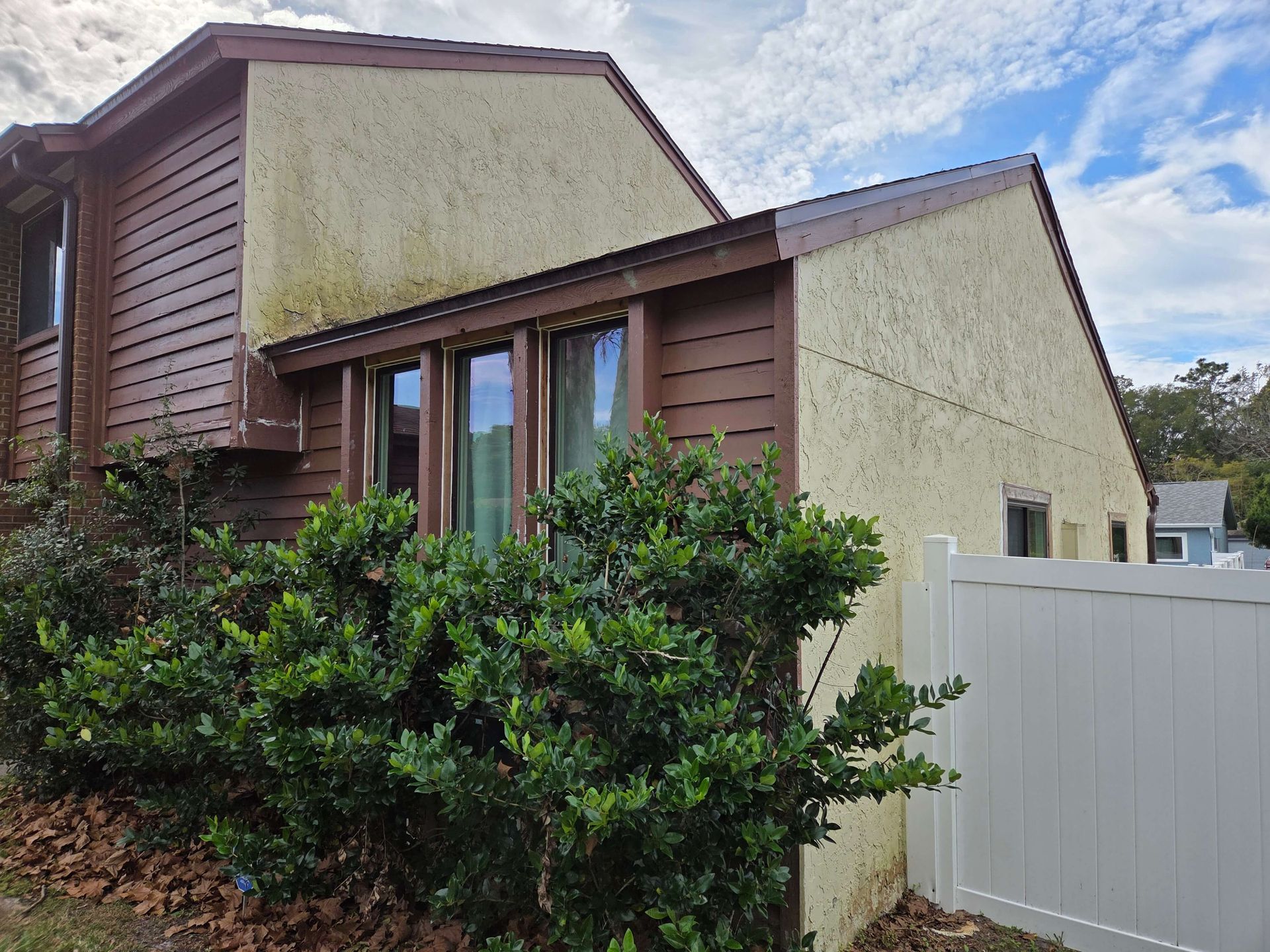Modern house exterior with brown and cream siding, large window, shrubs, and white fence under a cloudy sky