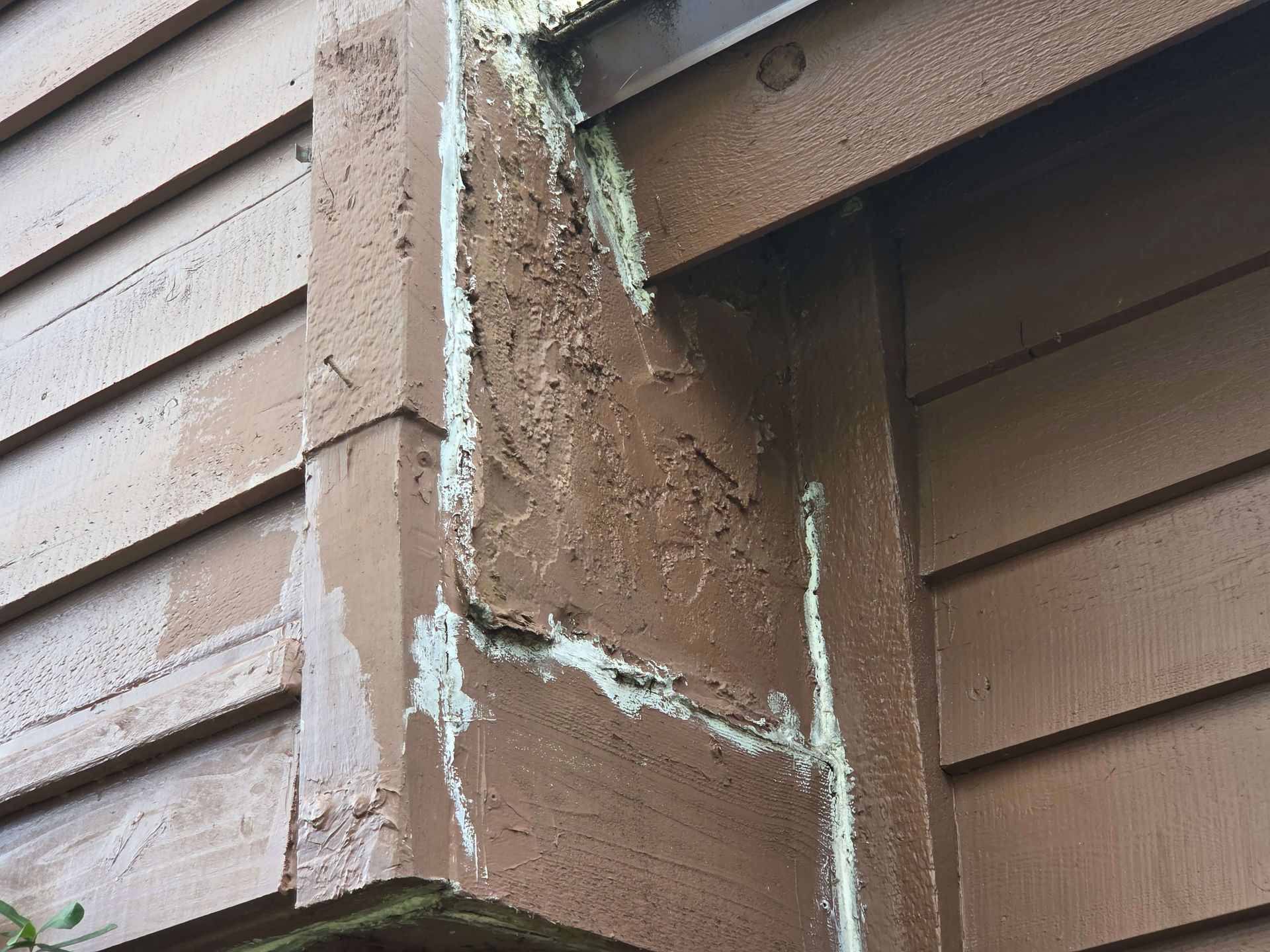 Close-up of damaged brown wood siding with peeling white sealant at a corner seam