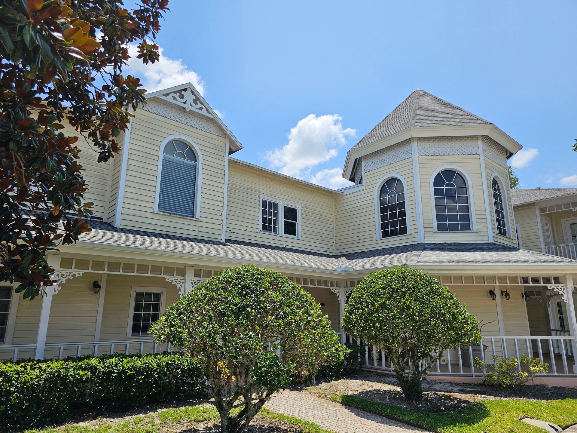 Two-story cream house with white trim, curved porch, and trimmed shrubs under a blue sky.