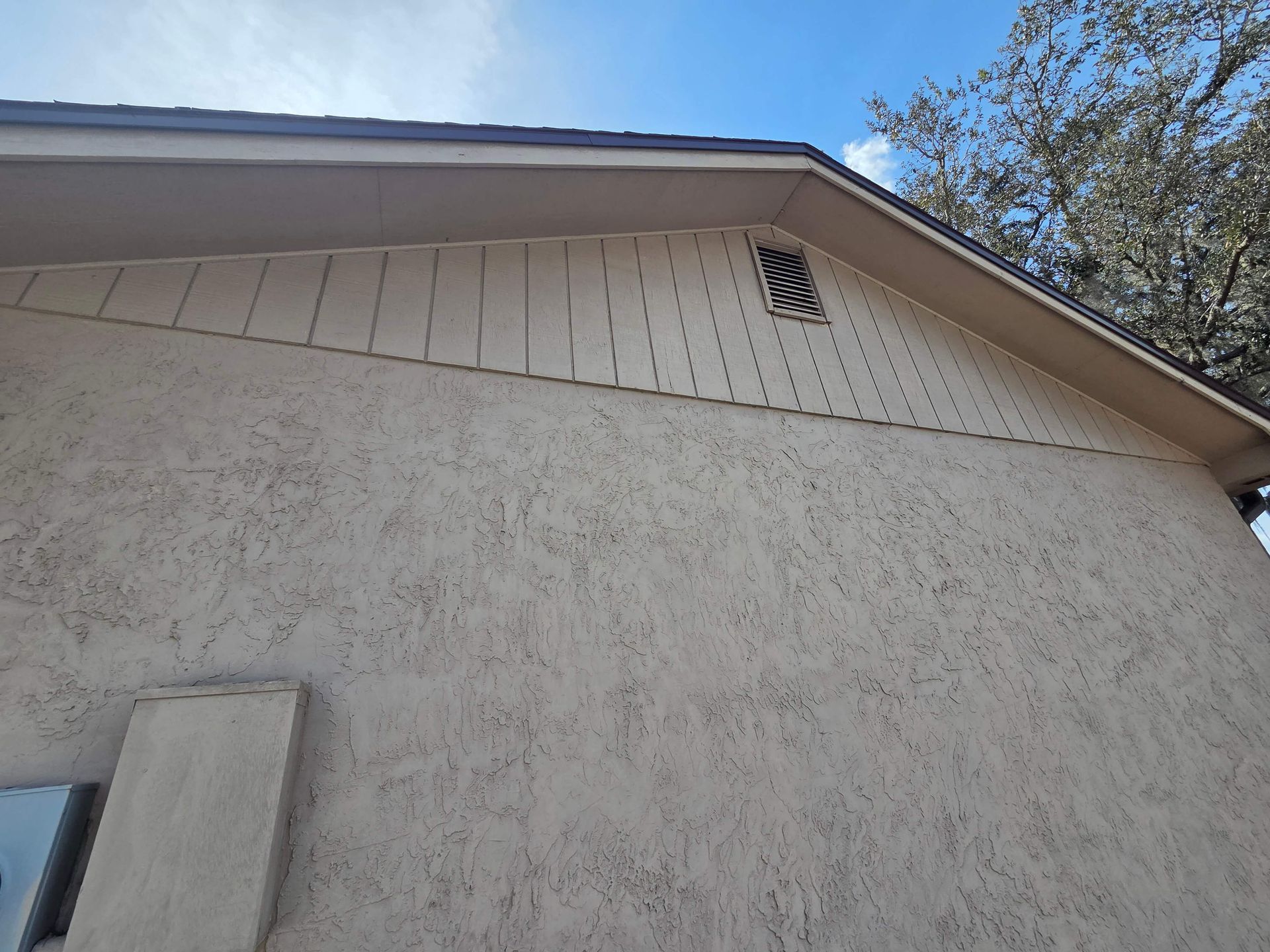Exterior view of a beige stucco house with gable roof, eaves, and a small vent under a blue sky