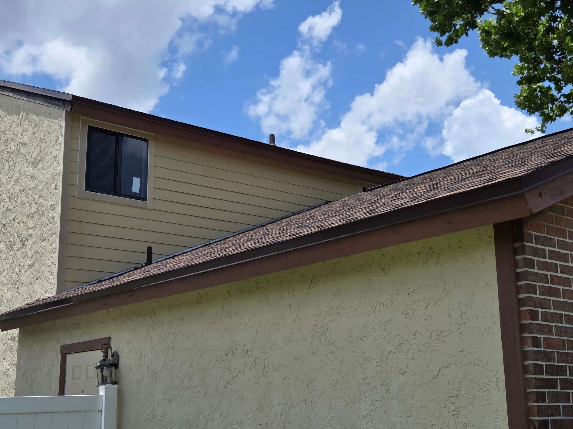 Exterior of a house with beige siding, a sloped brown roof, and blue sky with clouds above