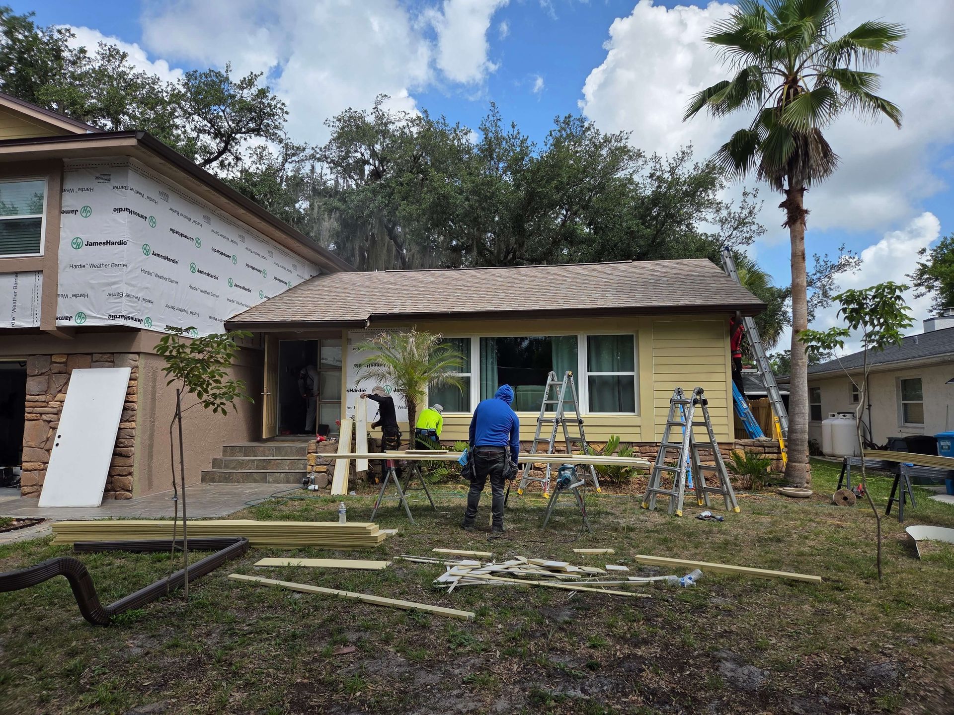 Workers install windows on a small beige house with ladders and construction materials in the yard