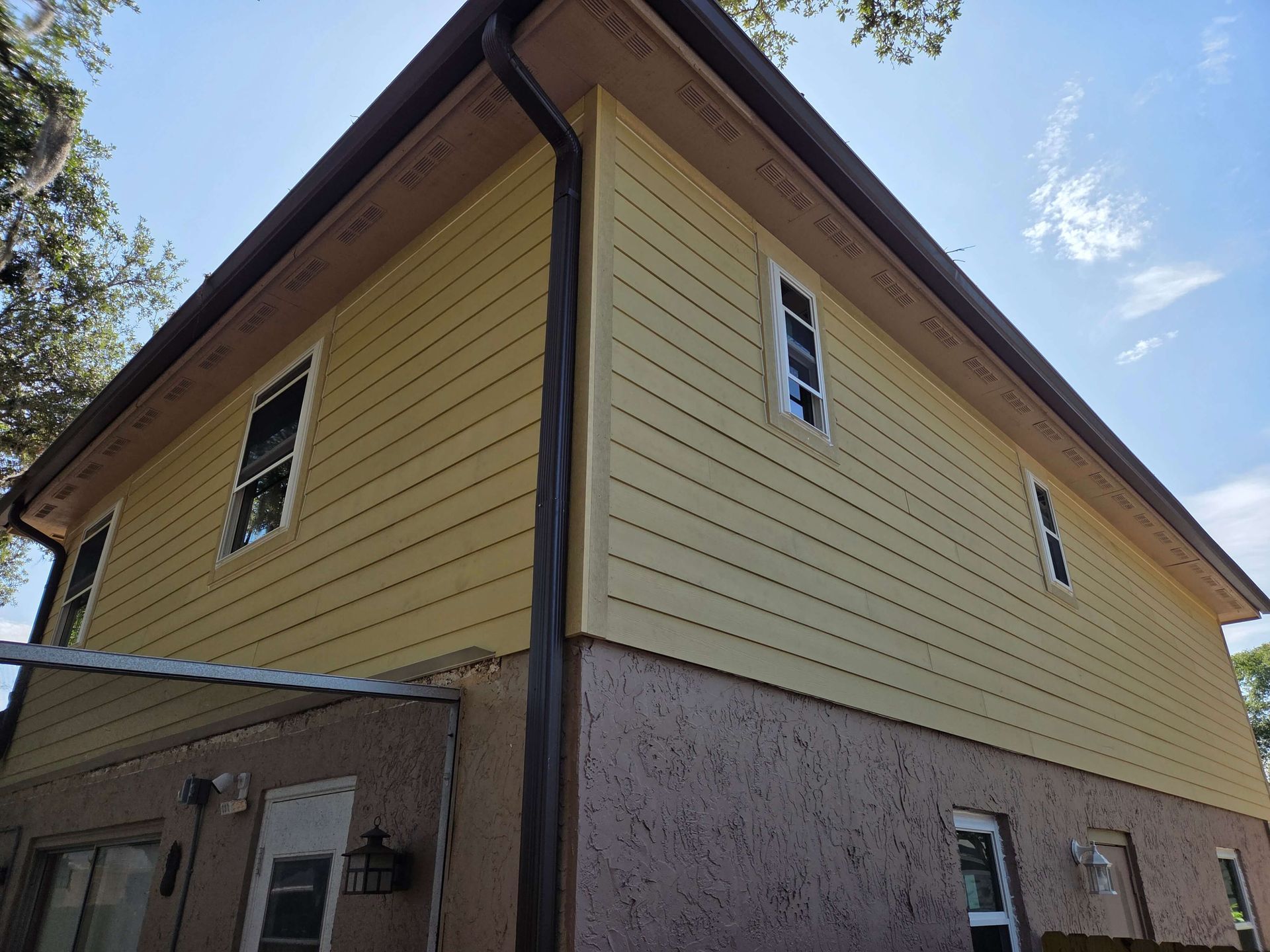 Two-story beige house with brown trim and stone lower exterior, viewed from below at the corner.