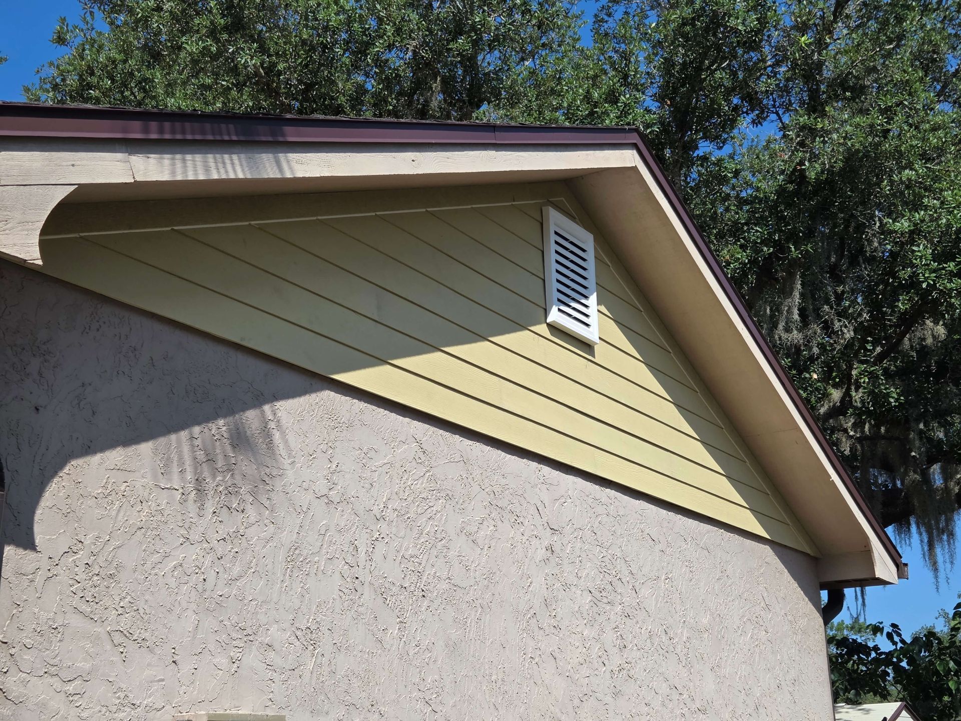 House exterior with beige siding, white vent, and sloped roof under trees