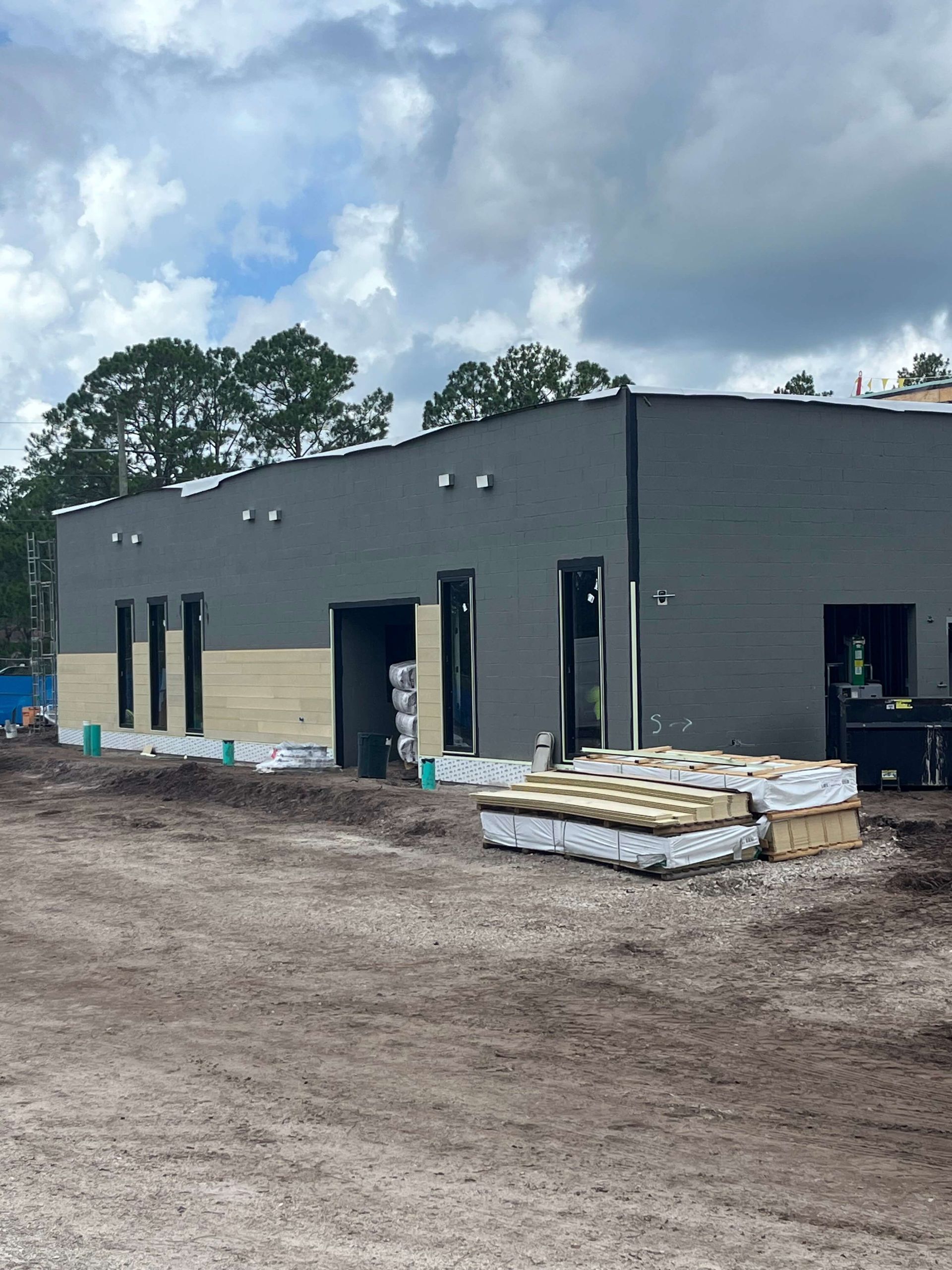 Gray commercial building under construction with black doors and windows, framed by dirt and stacked materials
