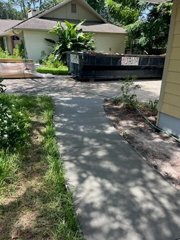 Concrete walkway beside a house leading to a black dumpster in a sunny yard