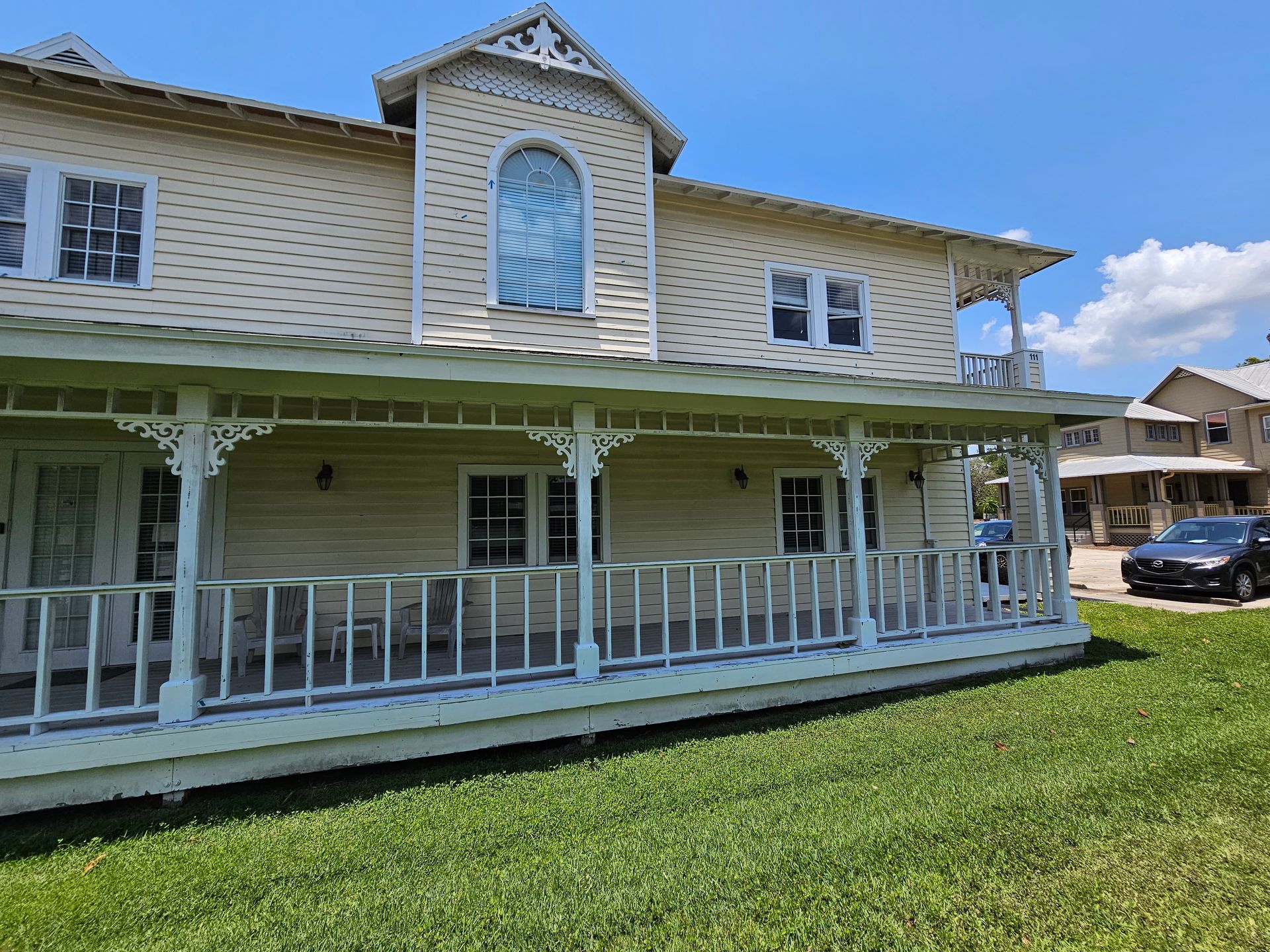 Two-story white house with a front porch, green lawn, and a parked car beside it under a blue sky