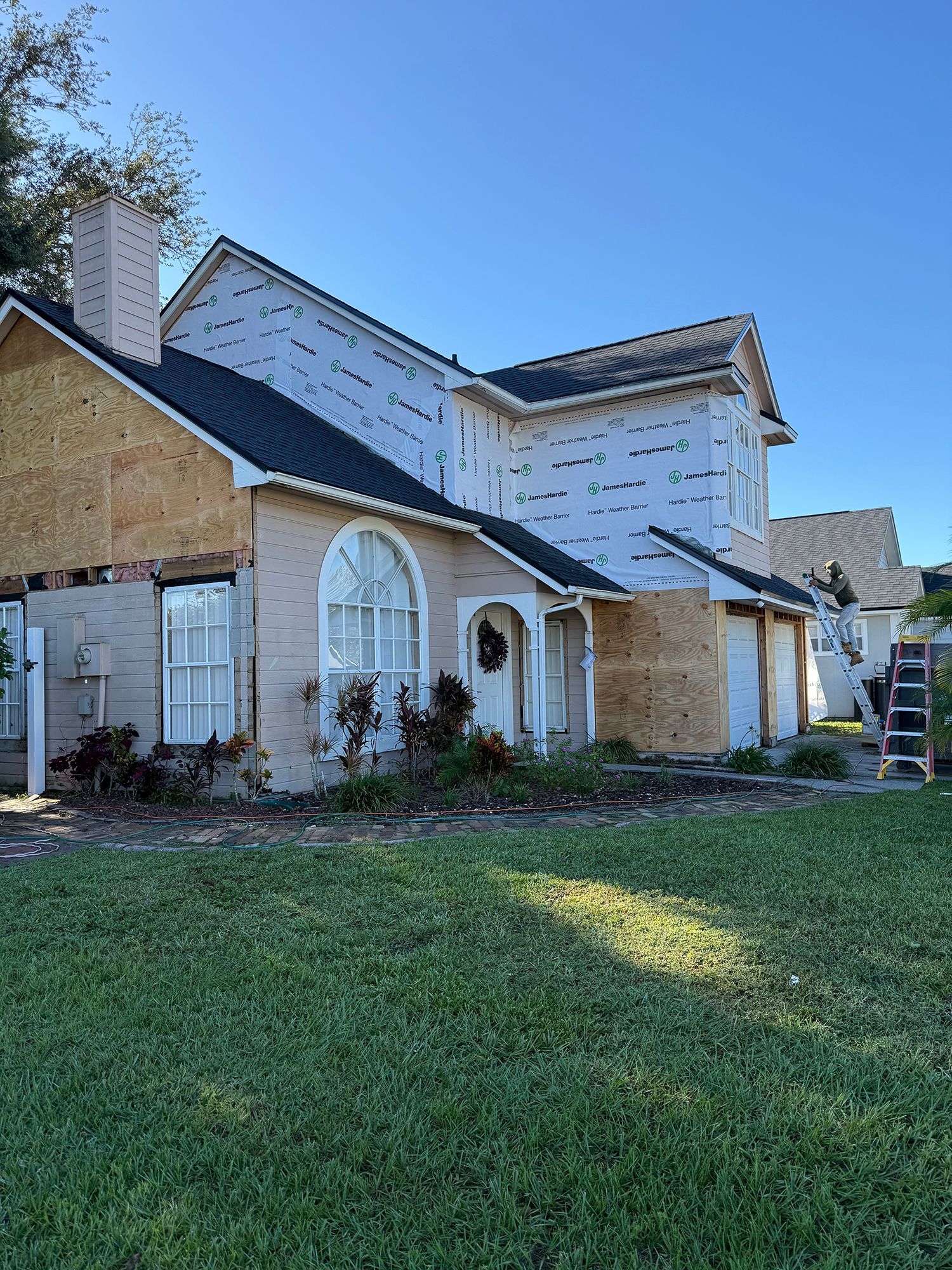 Suburban house exterior with tan brick, white siding, and a green lawn under a clear blue sky